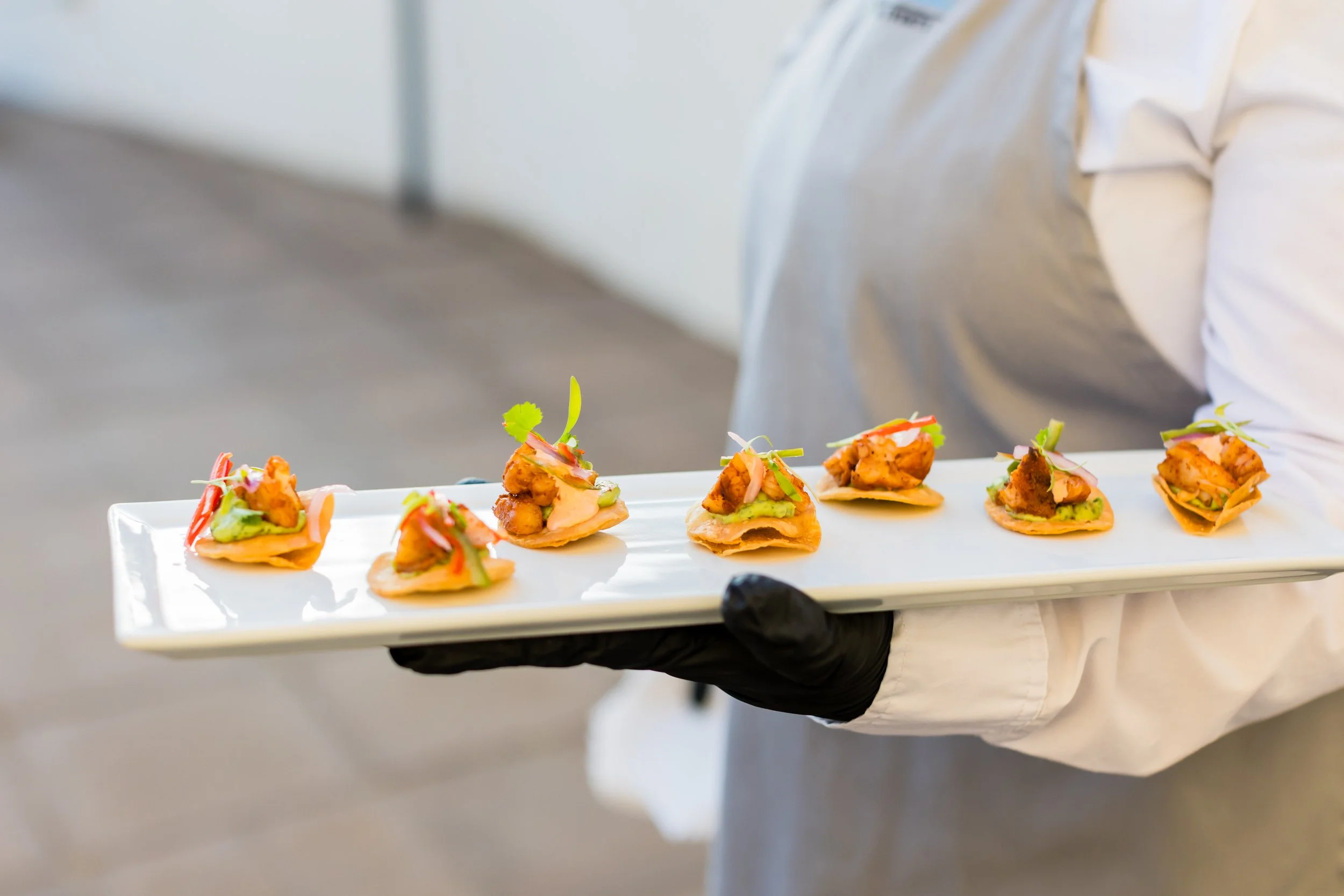 Person holding a white tray with six small gourmet appetizers, garnished with greens and sauces, in a professional setting.