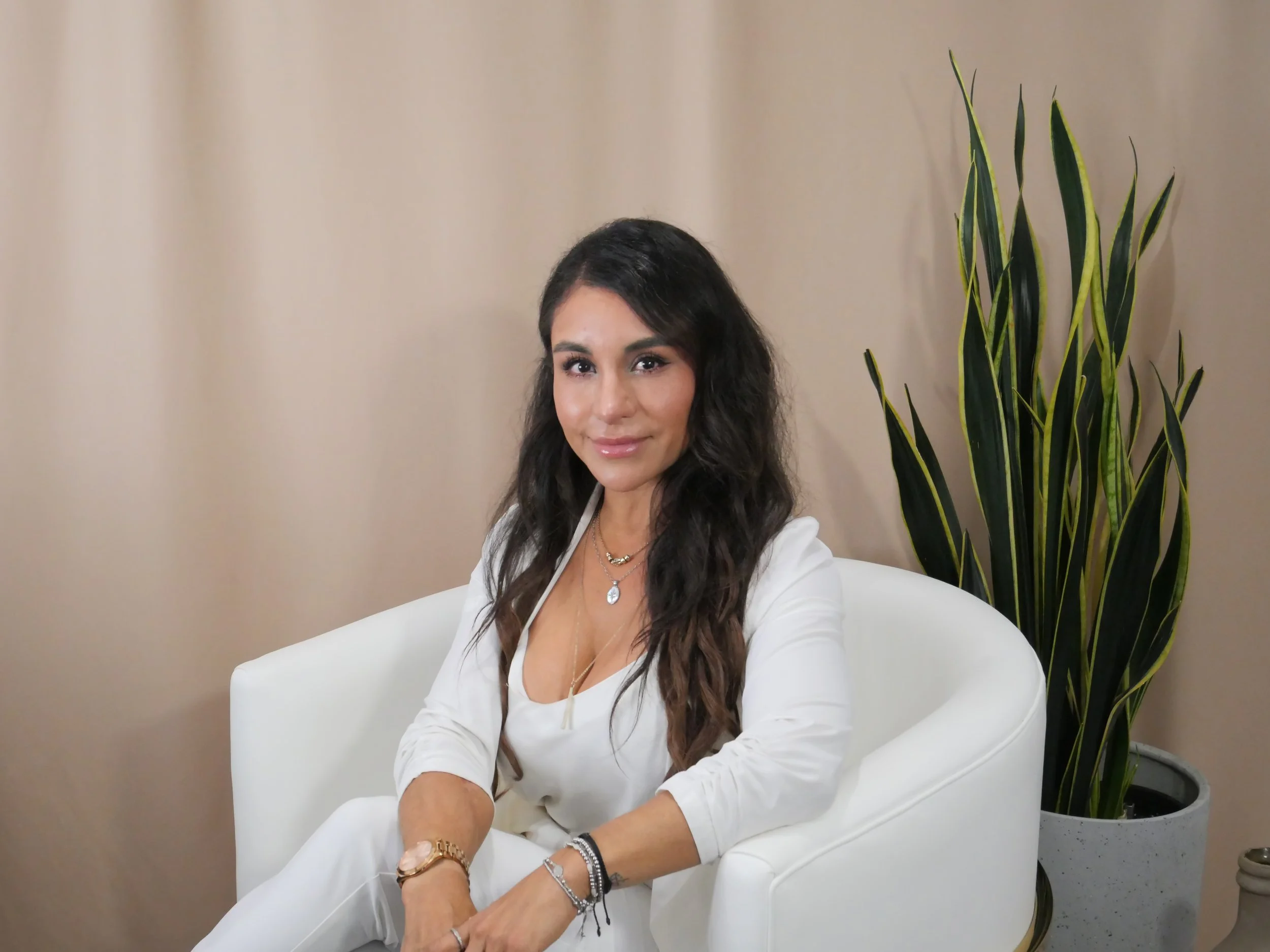 A woman with long, dark wavy hair sitting in a white armchair, wearing a white long-sleeve top and light-colored pants, with jewelry including necklaces and bracelets, next to a tall green plant in a pot, against a beige wall.