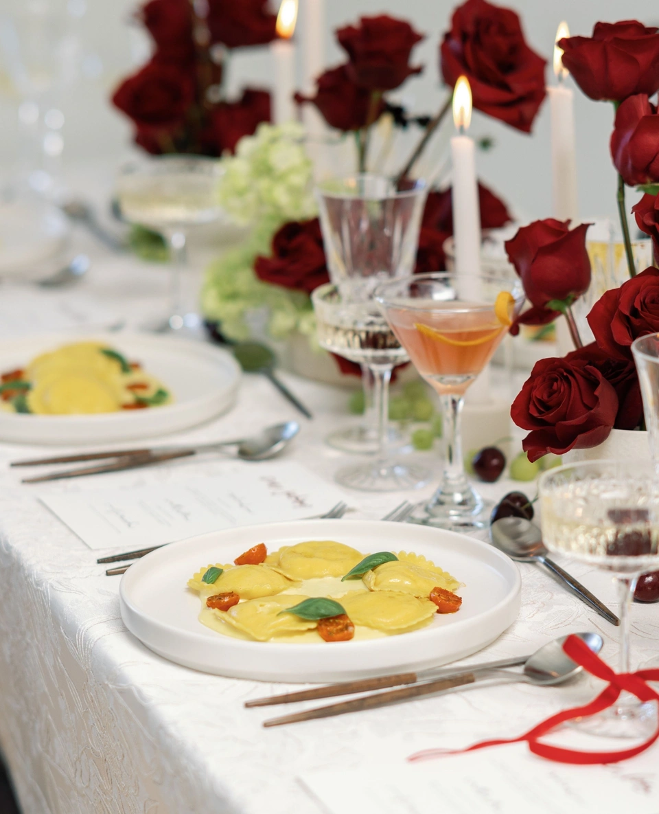 A formally set dinner table with a plate of ravioli garnished with basil and cherry tomatoes, topped with a creamy sauce, surrounded by wine glasses, candles, flowers, and utensils.