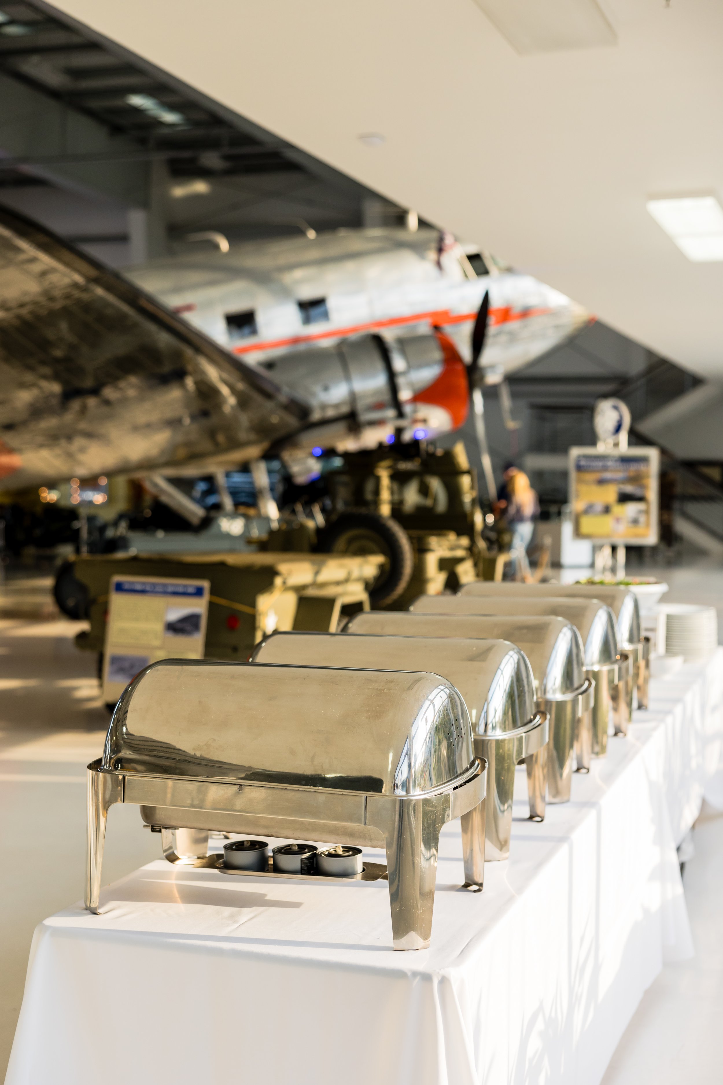 Buffet setup with chafing dishes on a table in front of a vintage airplane display.