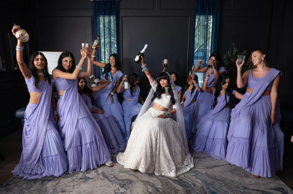 A group of women in purple traditional dresses celebrating indoors, holding bottles and glasses, with a bride in a white dress and veil in the center.