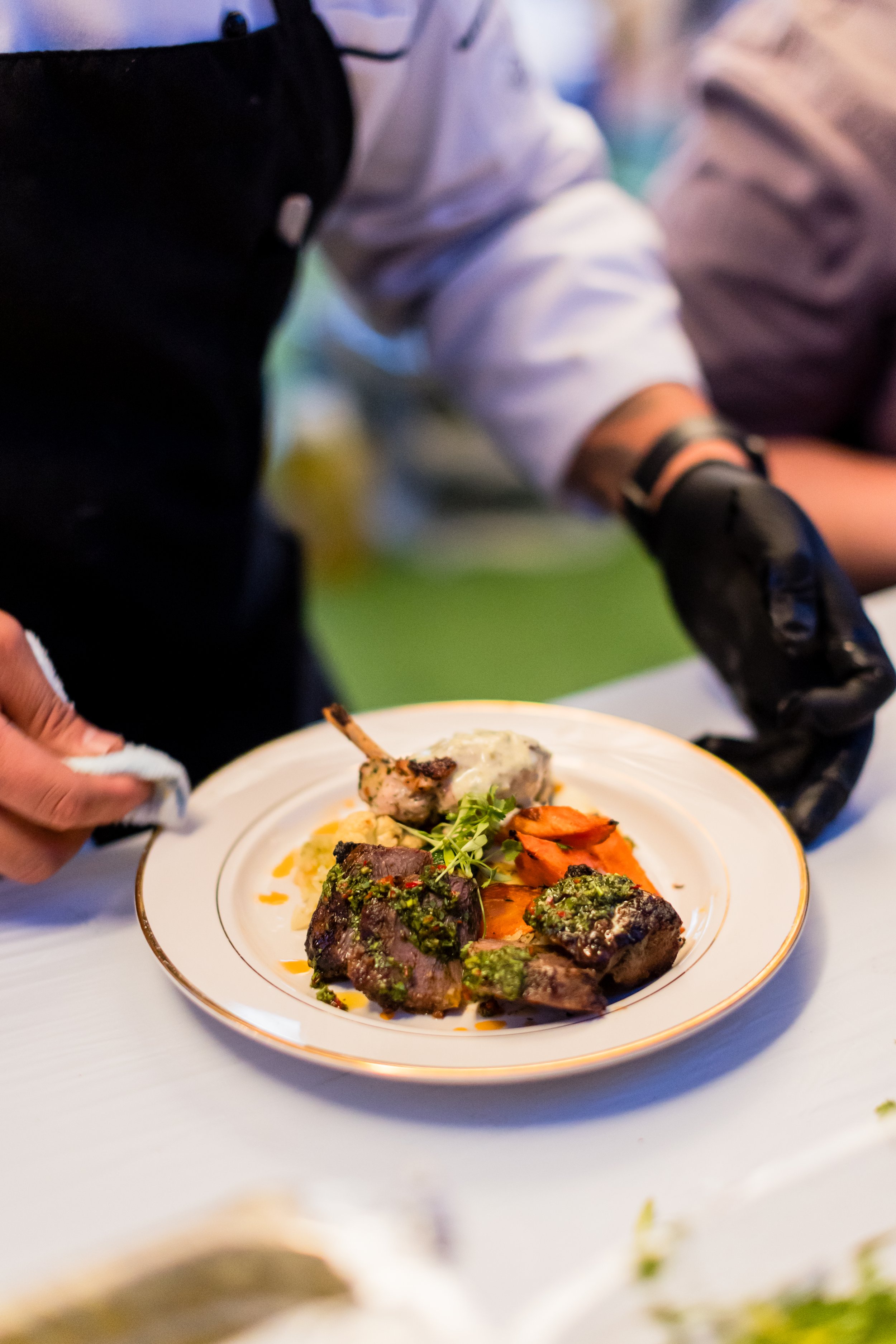Chef garnishing a plate with cooked meats and vegetables at a culinary event.