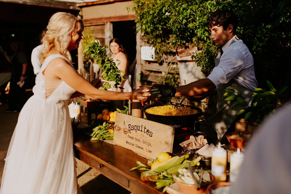 A woman in a white dress is receiving food from a man behind a food stand at an outdoor event. The stand has a sign listing items like fresh corn, cotija, Mexican cheese, and line. There are vegetables and condiments on the counter, and a woman is vi