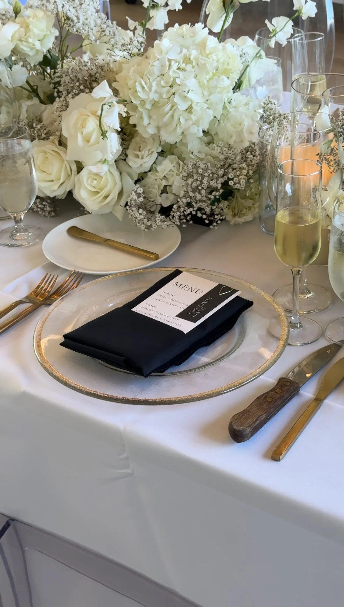 Elegant table setting with a white tablecloth, gold flatware, black napkin, menu, and large floral centerpiece of white roses, hydrangeas, and baby's breath, surrounded by champagne glasses and water glasses.