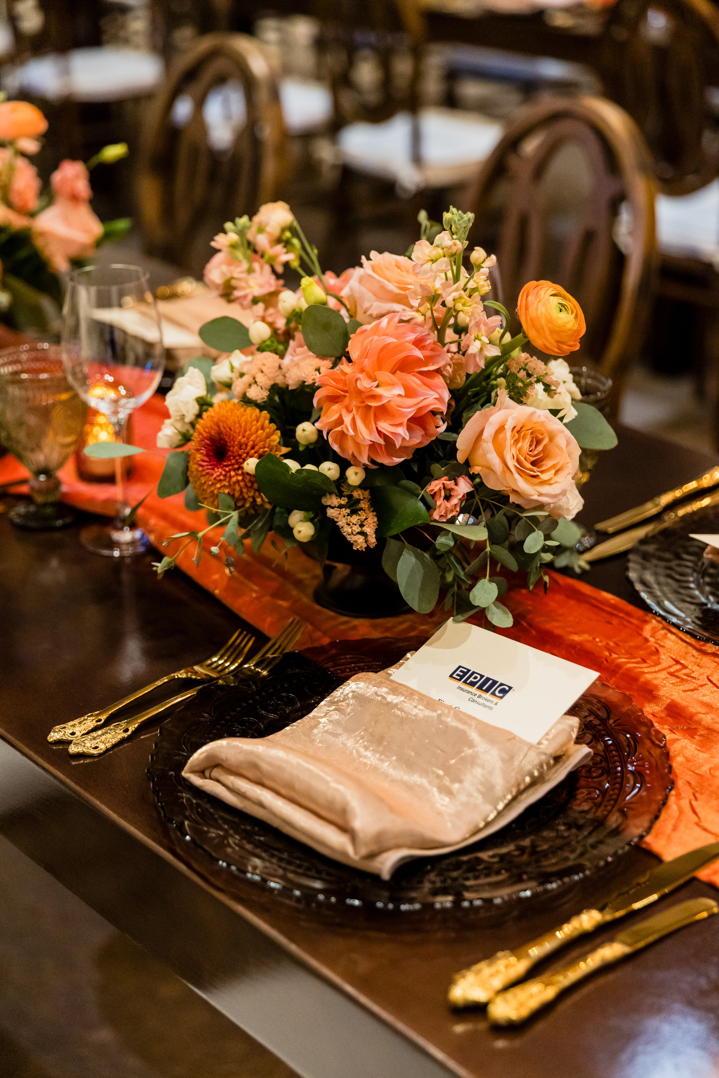 A formal dining table set for an event with a floral centerpiece, gold flatware, crystal glasses, and an orange table runner.