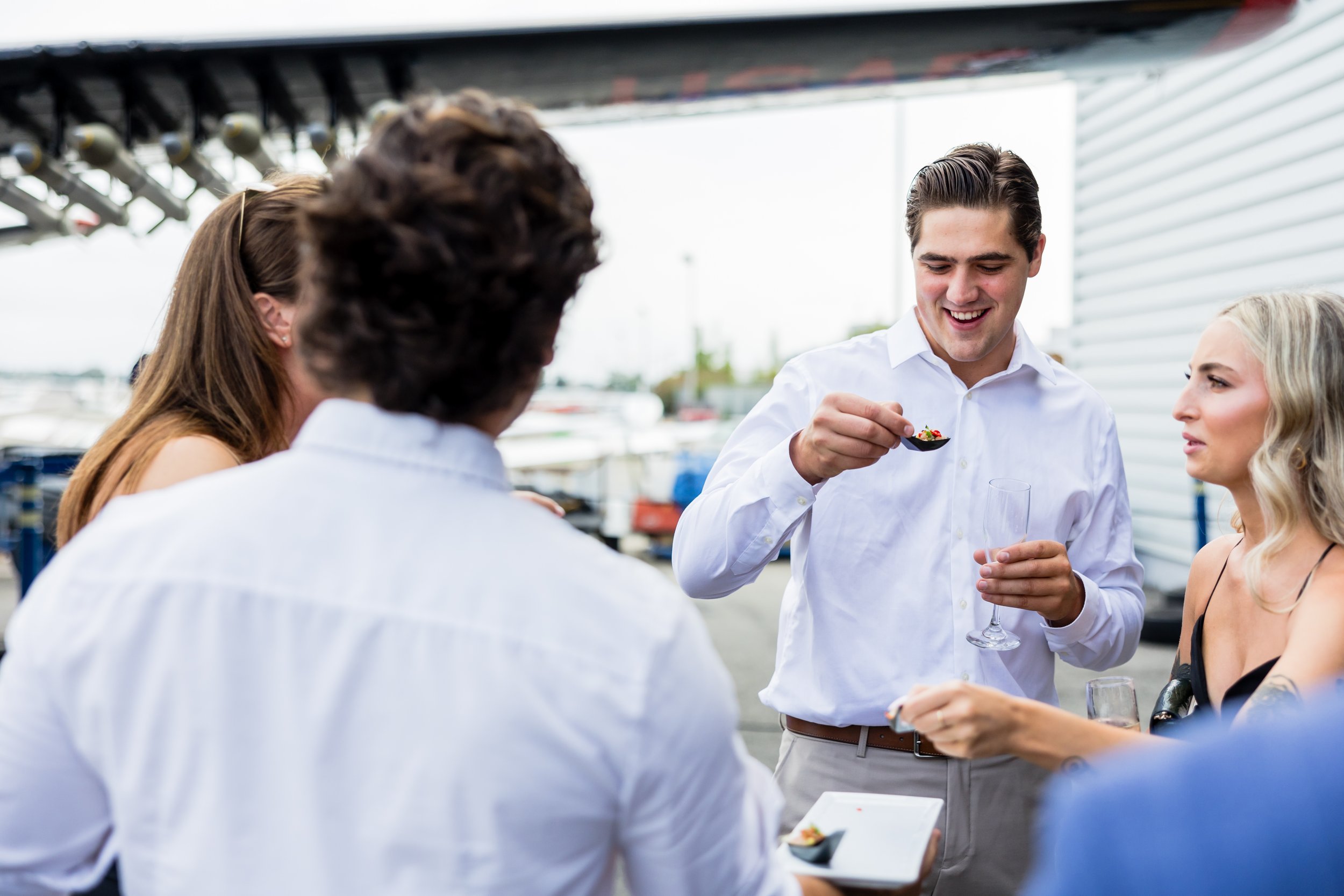 A group of four young adults socializing outdoors, with three of them in focus. One man is smiling while holding a spoonful of food and a champagne glass. Two women and one man are partially visible, one woman is holding a plate of food. They are dressed casually and appear to be enjoying a casual gathering.