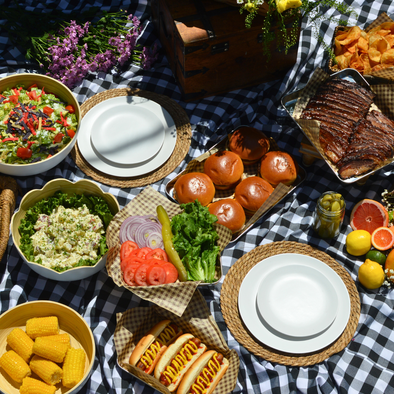 Picnic table set with a variety of foods including salad, bread rolls, ribs, hot dogs with mustard, potato salad, corn on the cob, and fresh fruit, all on a black and white checkered tablecloth.