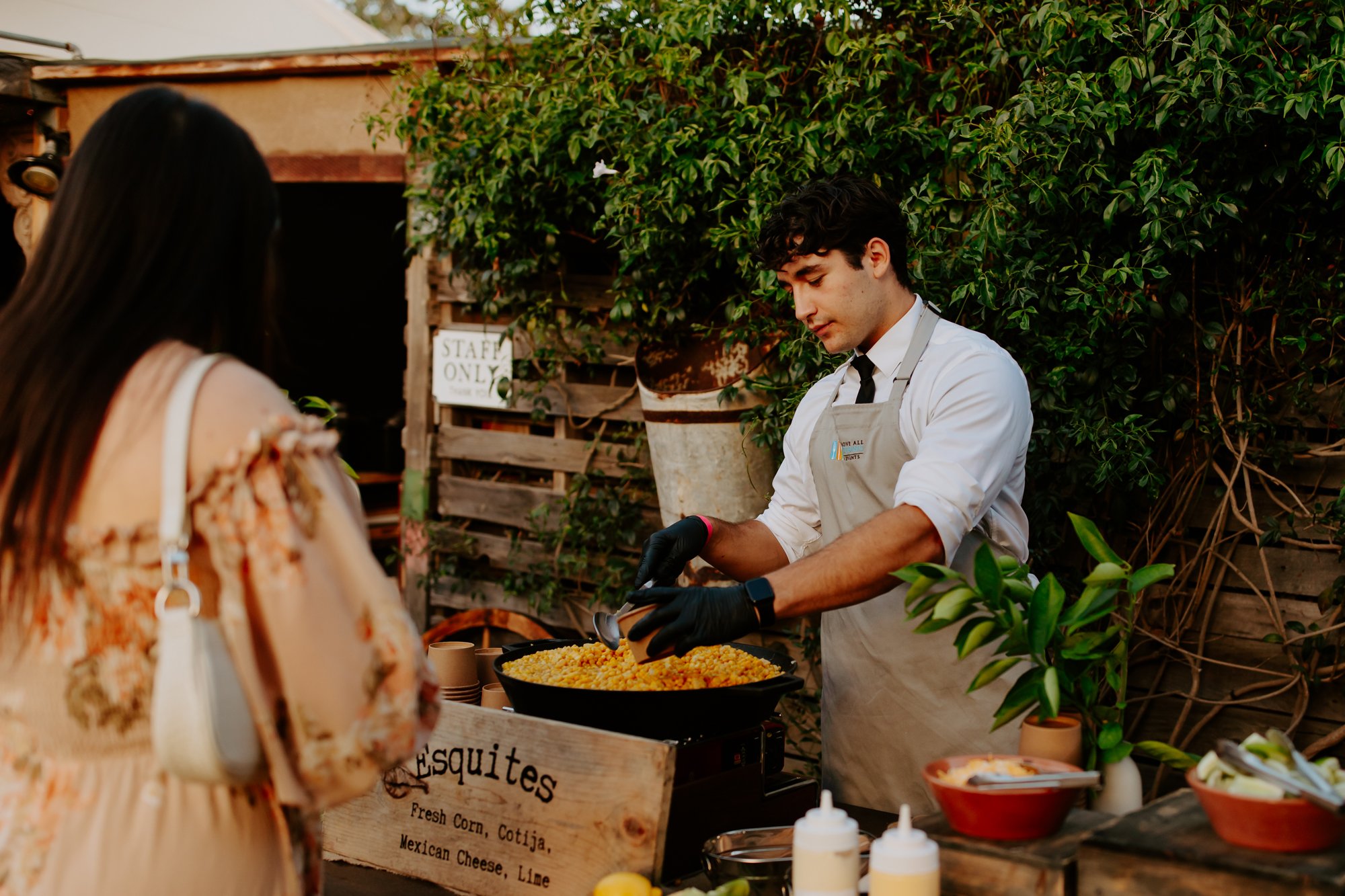 A young man serving food from a large pan at an outdoor food stand with a woman customer in front of him. The stand has a wooden sign with the words "Esquites" and ingredients listed. Plants and a wooden structure are in the background.