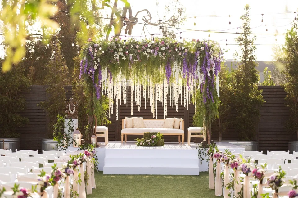 Outdoor wedding ceremony setup with floral decorations, a white stage with a beige sofa, and rows of chairs adorned with flowers, under hanging string lights.