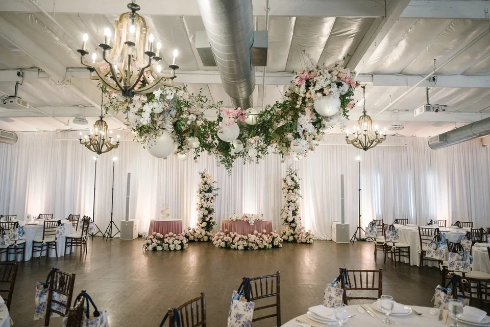 Elegant event space decorated with hanging chandeliers, floral arrangements, and draped white curtains, set up for a wedding reception or celebration.