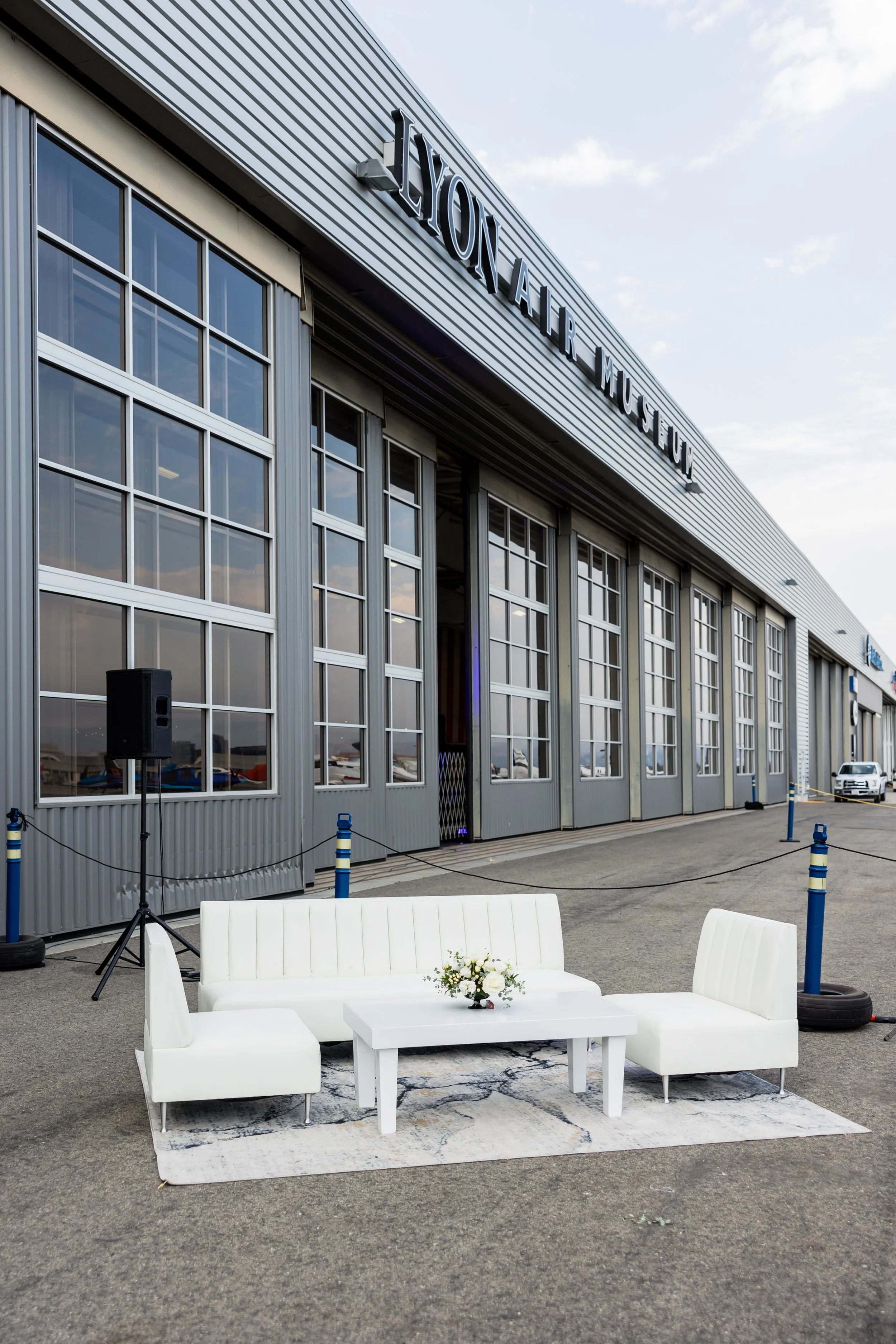 Outside view of a building labeled 'Air & Space Museum' with large windows. In front, there is a white sofa and two white armchairs surrounding a white coffee table with a plant. Blue posts with ropes cordon off the area, and a few cars are visible i