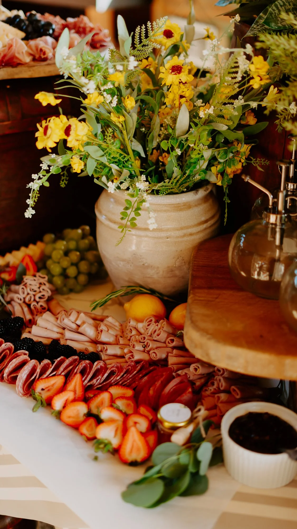 A floral arrangement with yellow flowers in a beige pot, surrounded by assorted fruits like strawberries, blackberries, white and green grapes, and lemons, along with sliced meats, napkins, and a small bowl of dark sauce on a wooden surface.