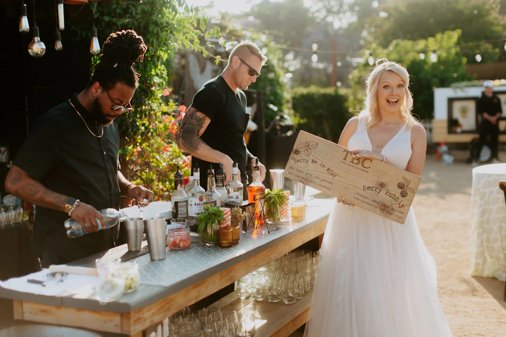 A woman in a white dress holding a wooden sign that says 'Berry Me in Mint' and 'Berry Bubble', smiling at an outdoor event. Two men behind a bar setup are preparing drinks, with bottles of alcohol, glasses, and garnishes on the table, in a sunlit ou