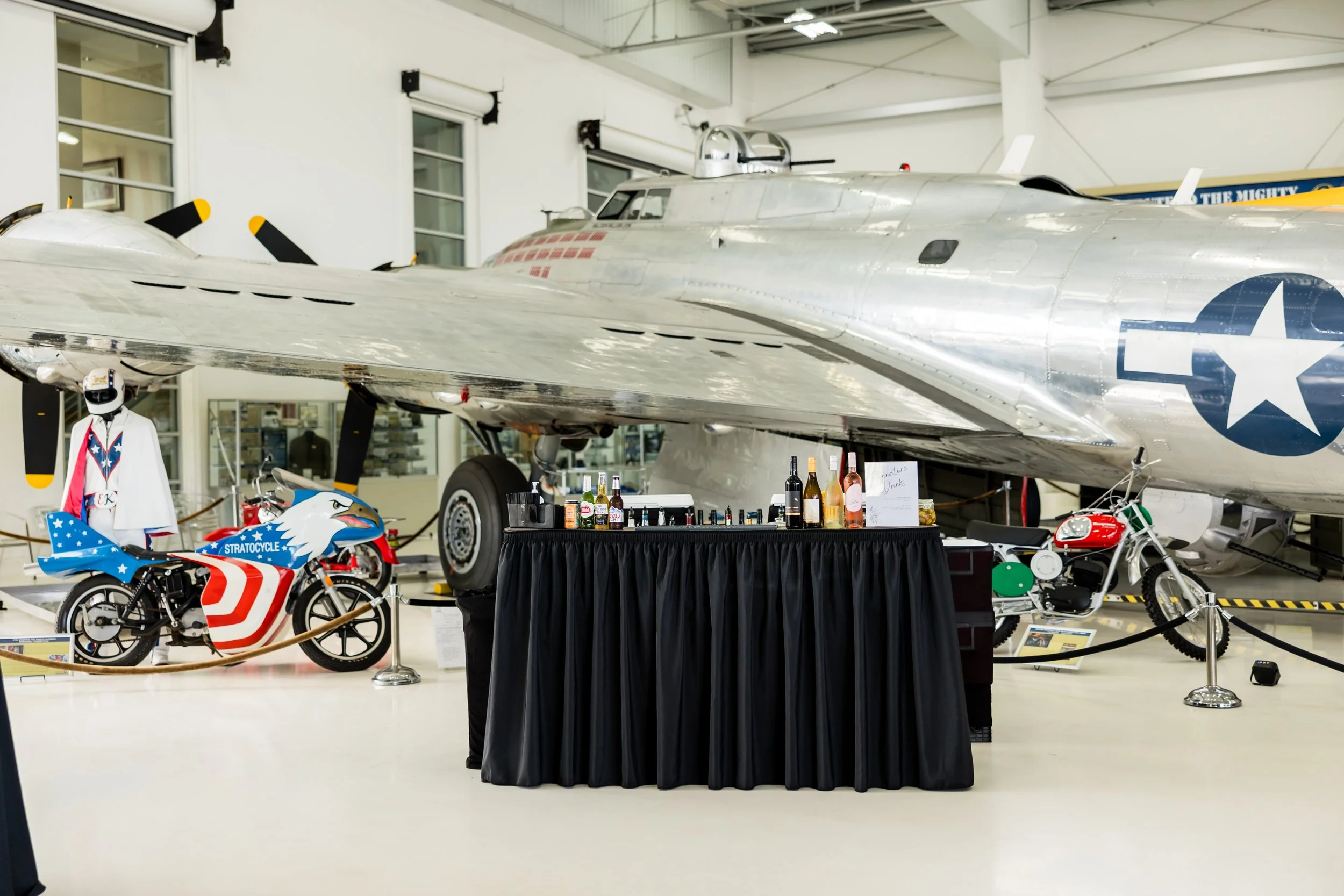 Vintage aircraft and motorcycles displayed in an indoor museum, with a bar setup in front of the aircraft.
