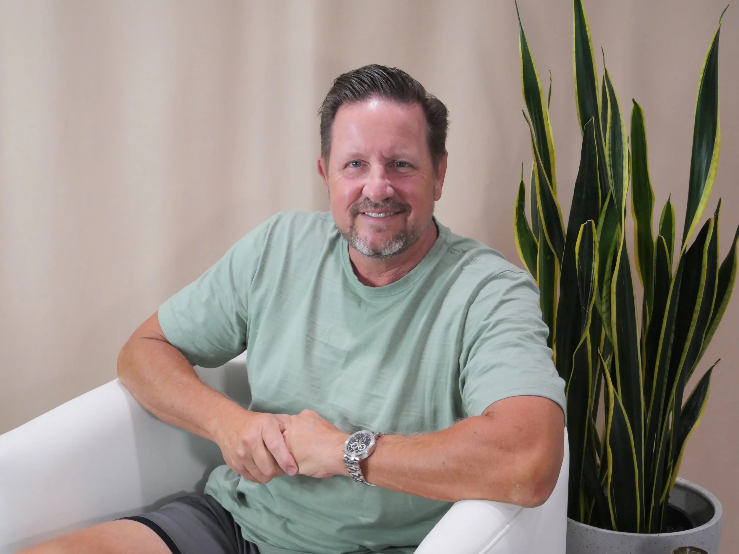 A man with short dark hair and a beard, wearing a light green t-shirt and a silver watch, sitting in a white chair next to a large green potted plant, smiling outdoors.