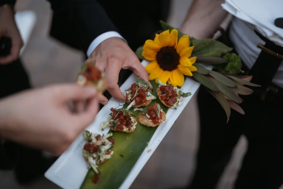 Person holding a tray of small appetizers garnished with red sauce, with a sunflower and greenery decoration.