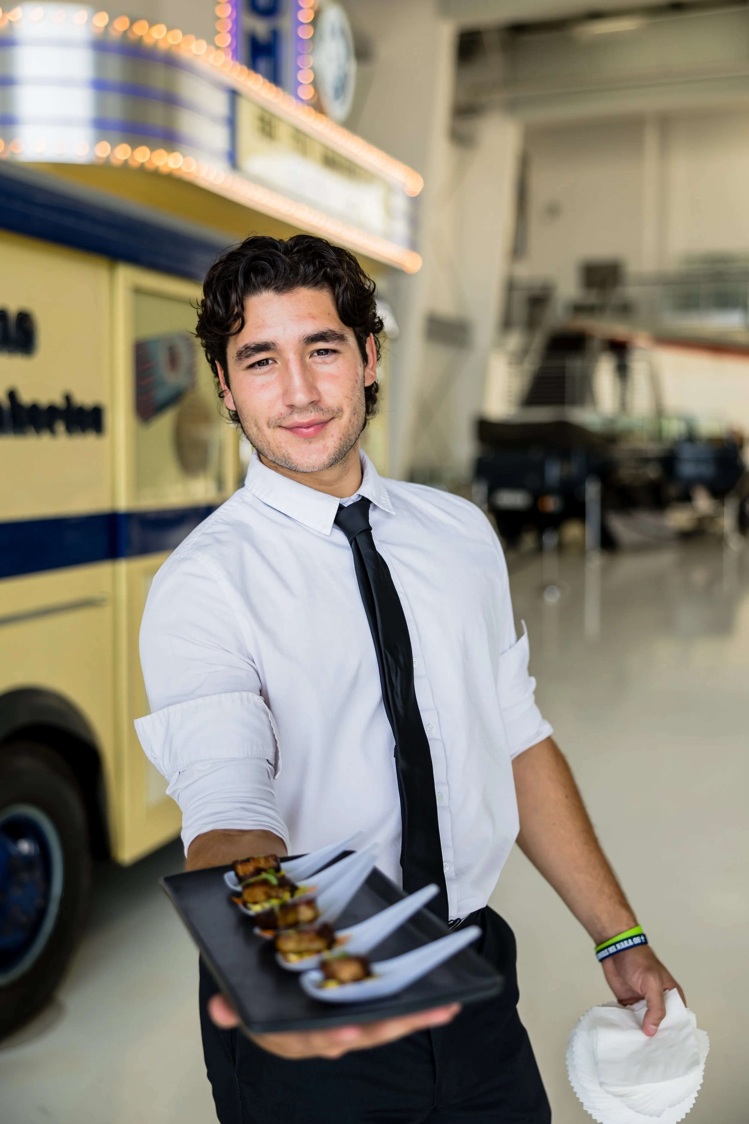 A young man in a white shirt with rolled-up sleeves, black tie, and a wristband, smiling and holding a tray with small servings of food on white spoons, standing inside a building with a yellow food truck and vintage vehicles in the background.