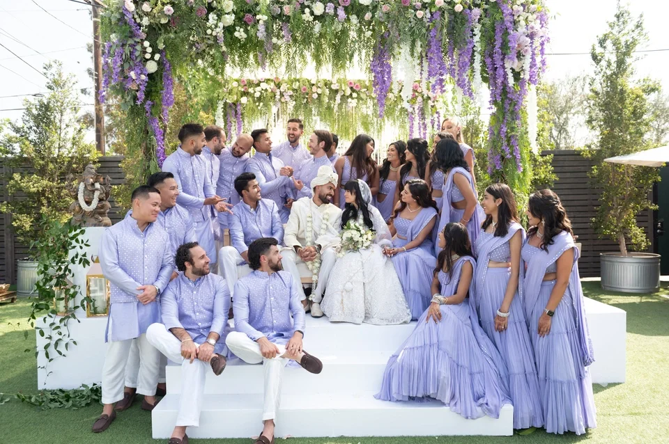 A wedding celebration outdoors with a large group of people, including the bride and groom, all dressed in light purple and white attire, gathered under a floral arch with purple and white flowers.