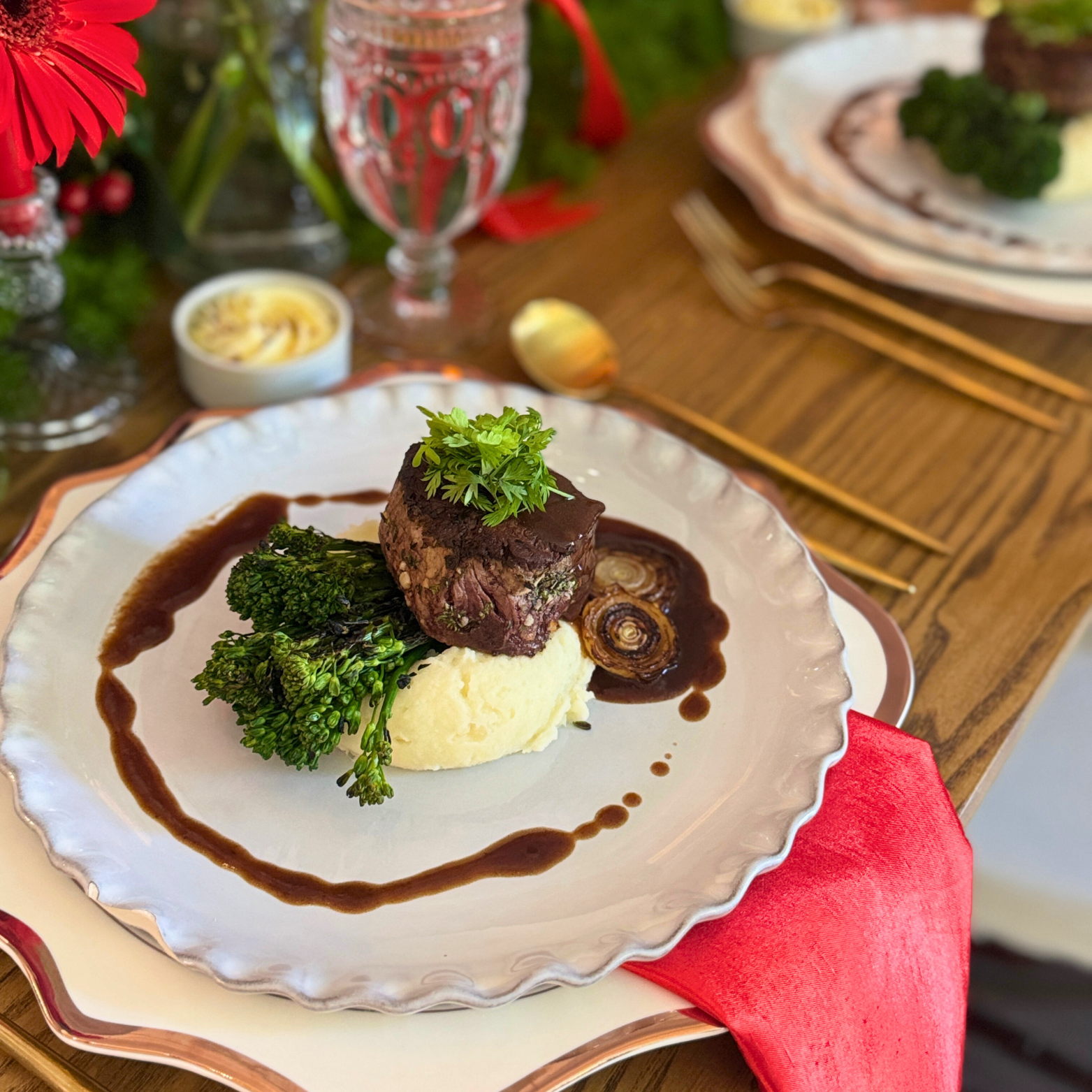 A plated filet mignon steak served with mashed potatoes, garnished with a sprig of parsley, alongside broccolini and caramelized onions, with a side of brown gravy.