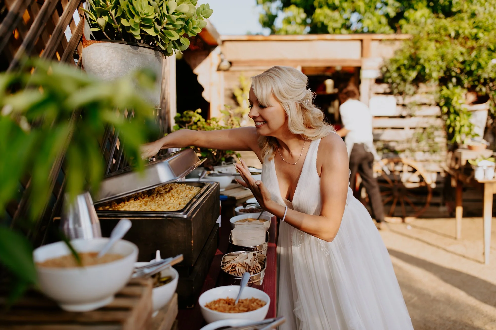 A woman dressed in a white wedding gown is serving herself food from a buffet table outdoors. The background features a rustic wooden setting and lush greenery.