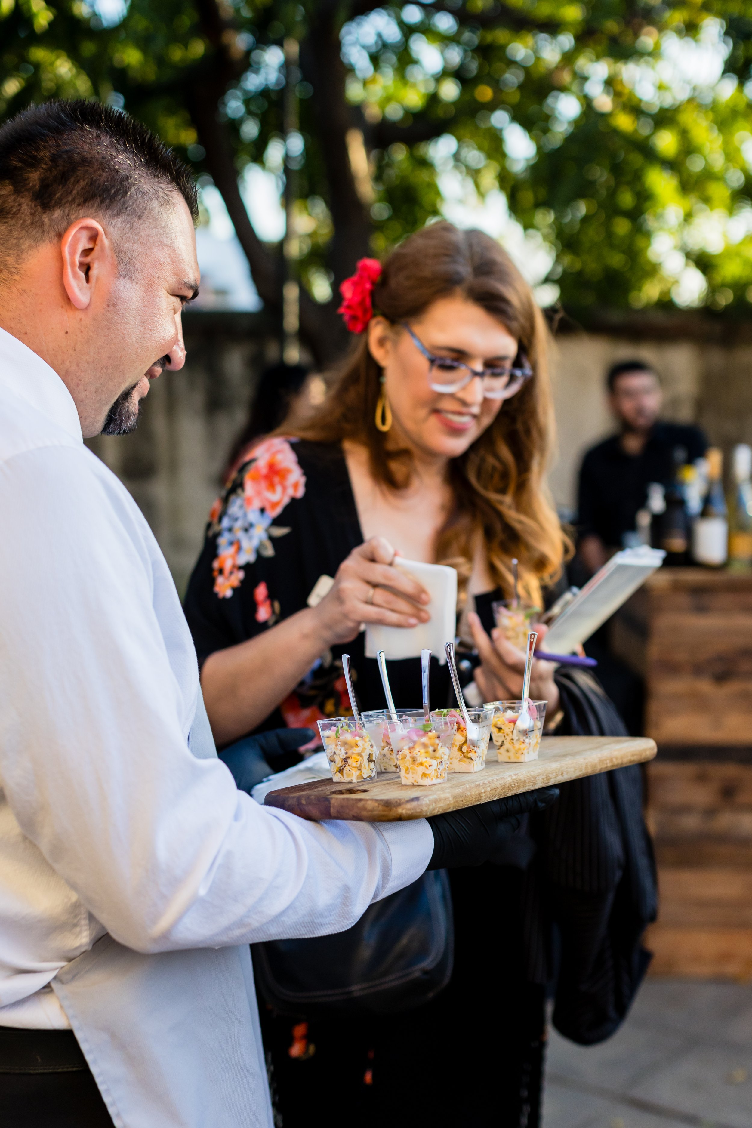 A waiter presenting a tray with four cups of salad at an outdoor event, with a woman in floral top holding a notebook in the background.