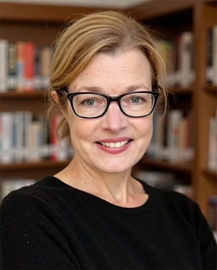 A woman with blonde hair, glasses, and a black shirt standing in front of bookshelves in a library.
