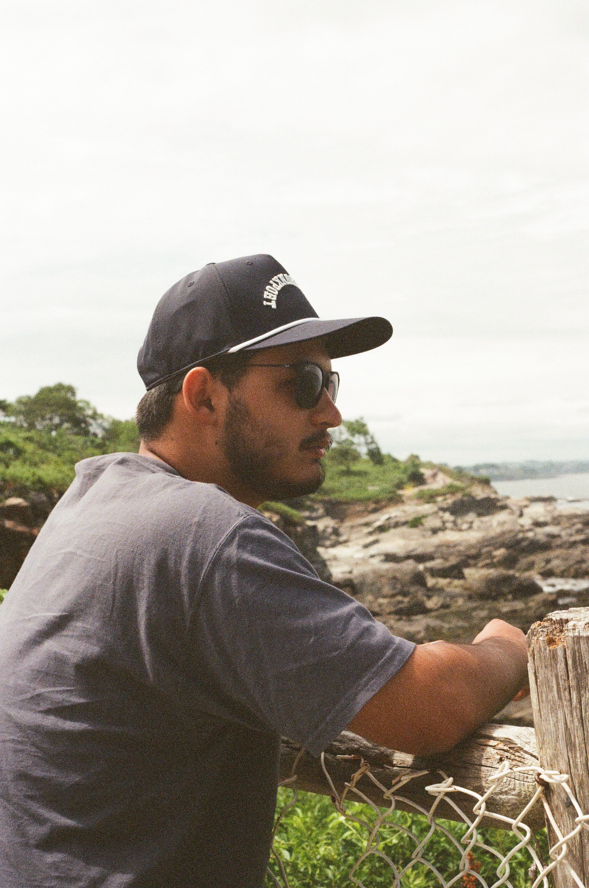 A man wearing a baseball cap, sunglasses, and a t-shirt leans on a wooden fence near a rocky waterfront, with trees and cloudy sky in the background.