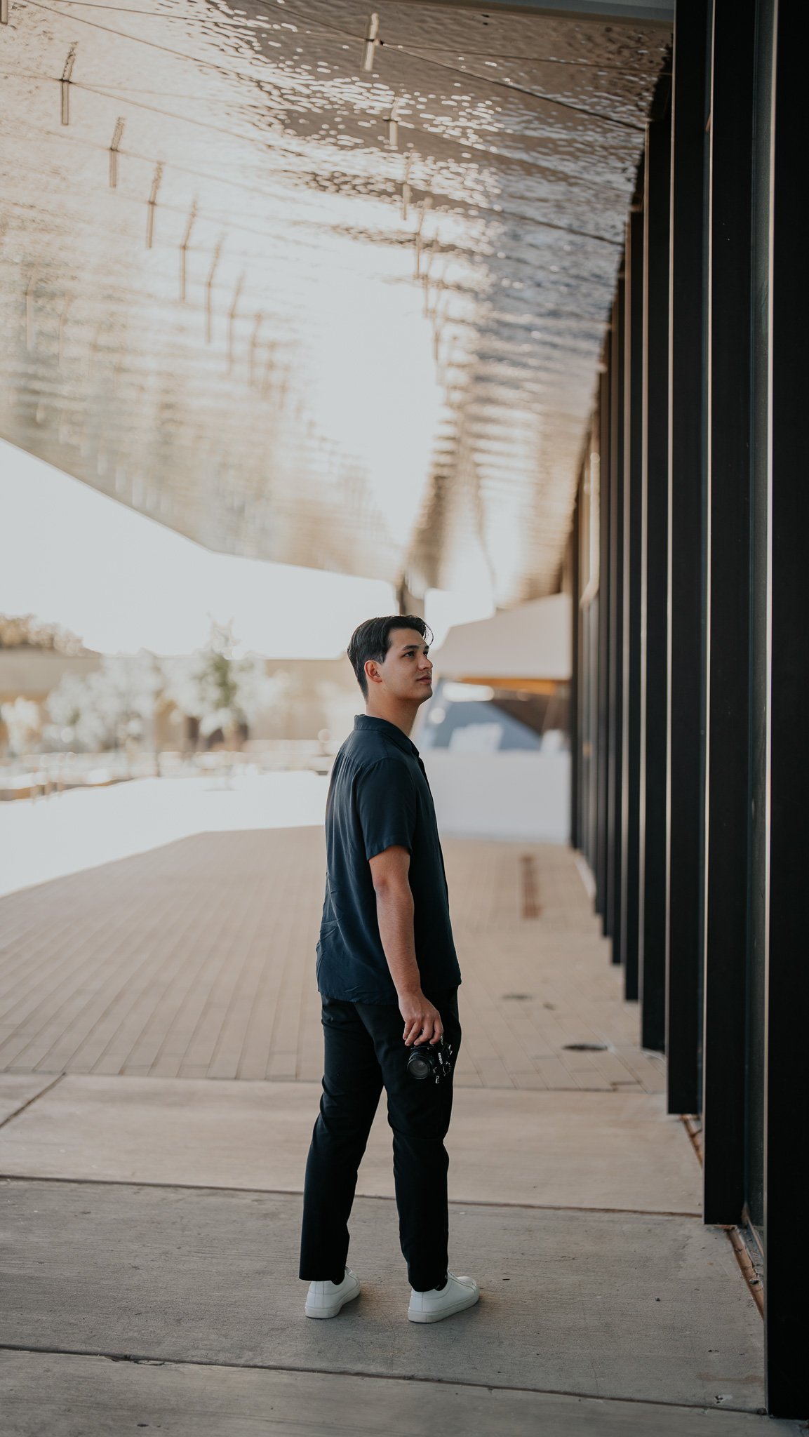 A man stands outdoors holding a camera, looking to his right, near a modern building with a metallic overhang and black support beams, during daytime with bright sunlight.
