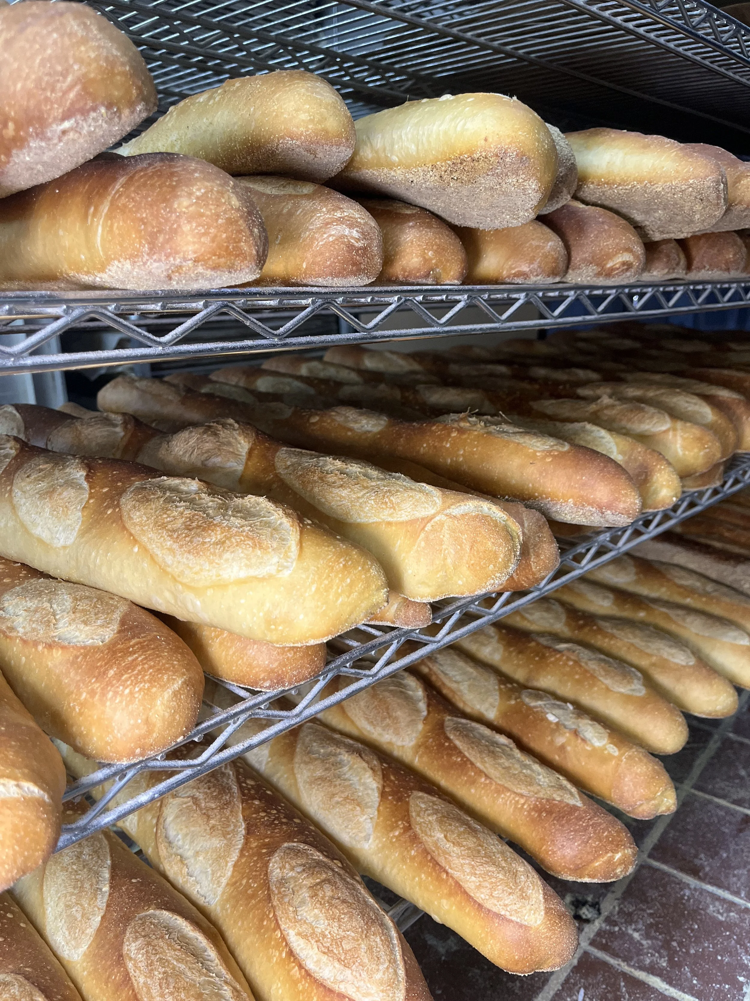 Multiple loaves of bread on metal racks in a bakery. The top shelf has rounded bread rolls, the middle and bottom shelves have elongated baguette-style loaves.