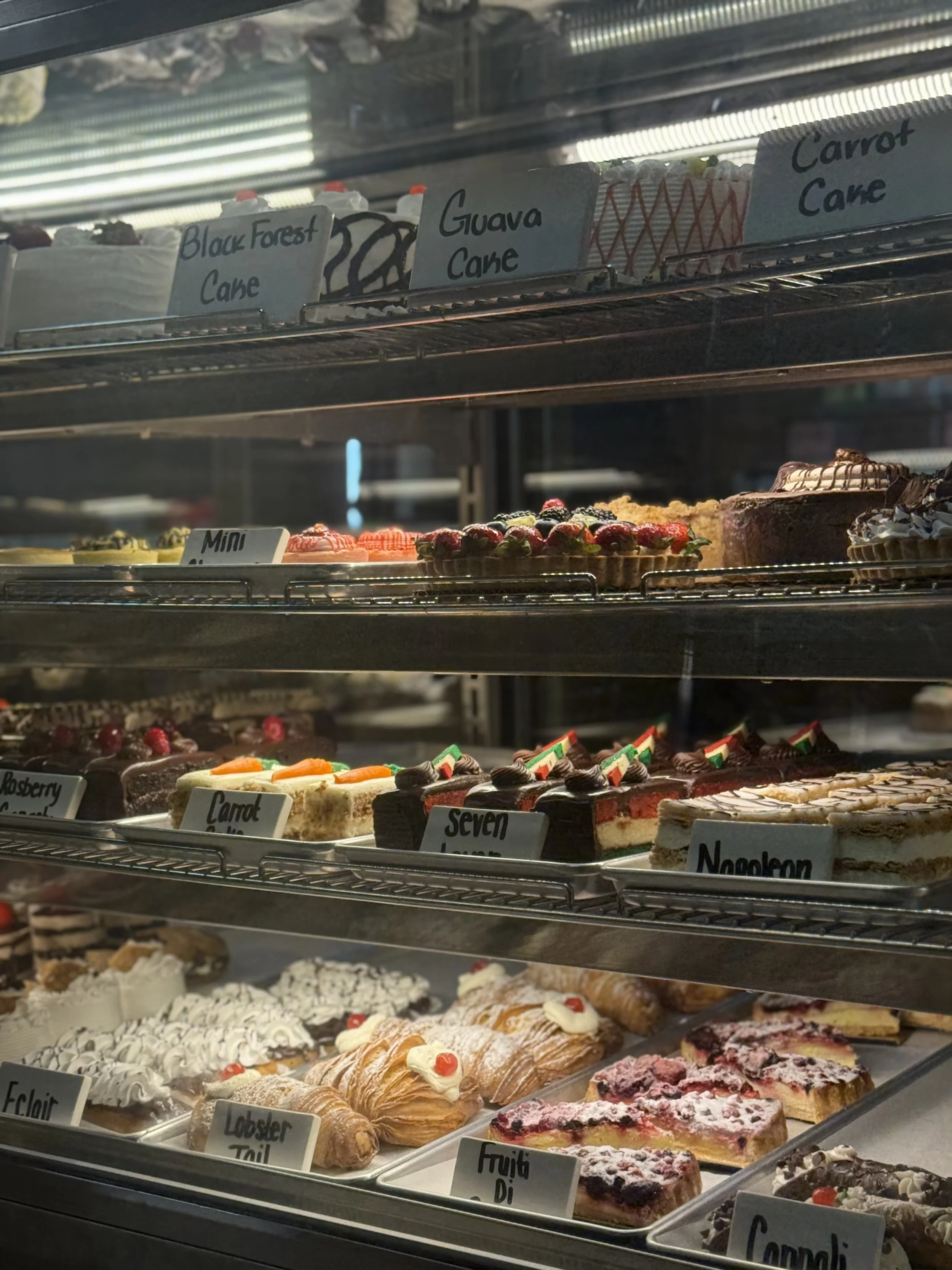 Display case filled with various pastries and cakes, including fruit tarts, eclairs, and layered cakes, labeled with names like Black Forest Cake, Guava Cake, Carrot Cake, and others.