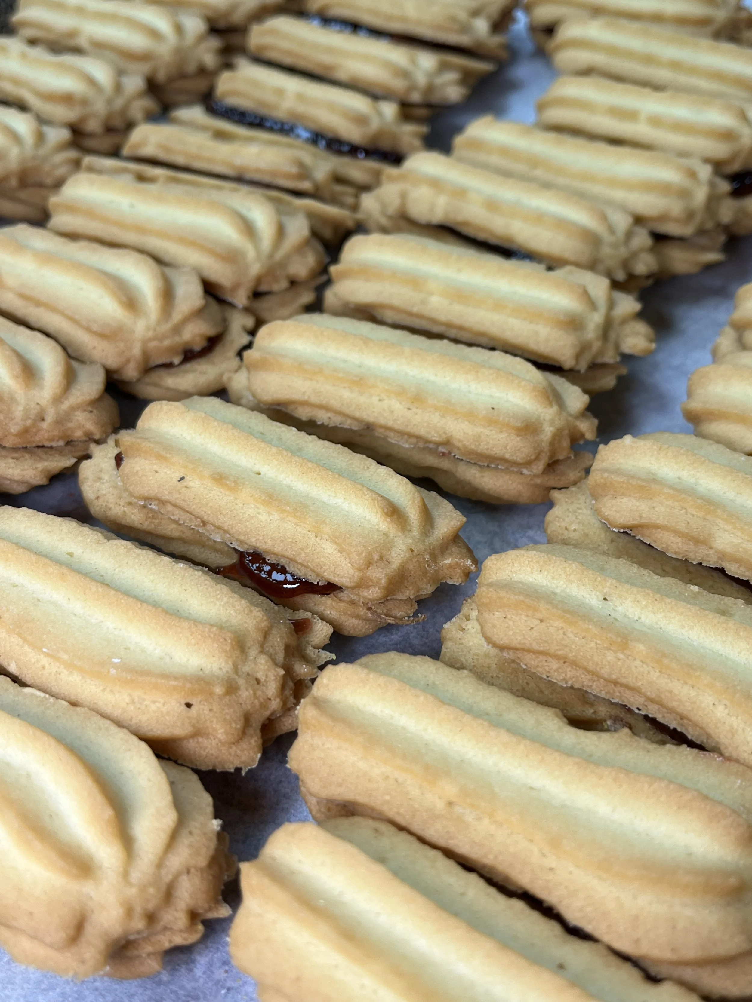 Close-up of numerous shortbread cookies filled with jam, arranged on parchment paper.
