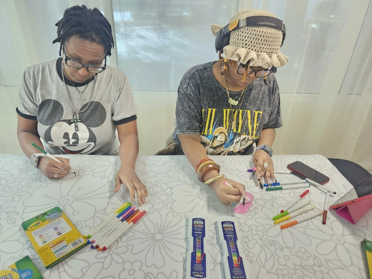 Two women coloring on a floral patterned table with colored markers, a pink tablet, and two color change markers.