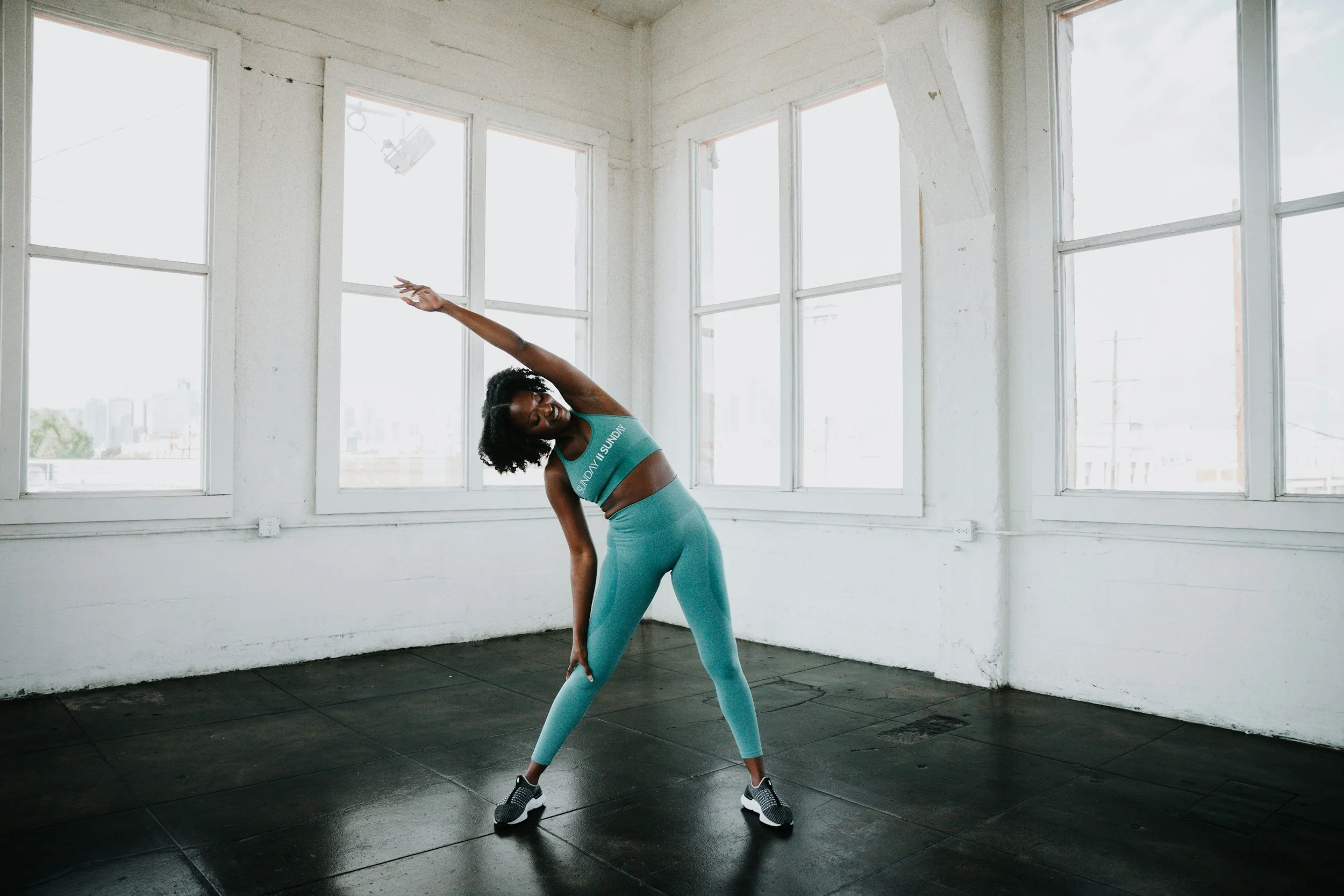 A woman wearing teal workout clothes stretched in a spacious, brightly lit studio with large windows and a black tiled floor.