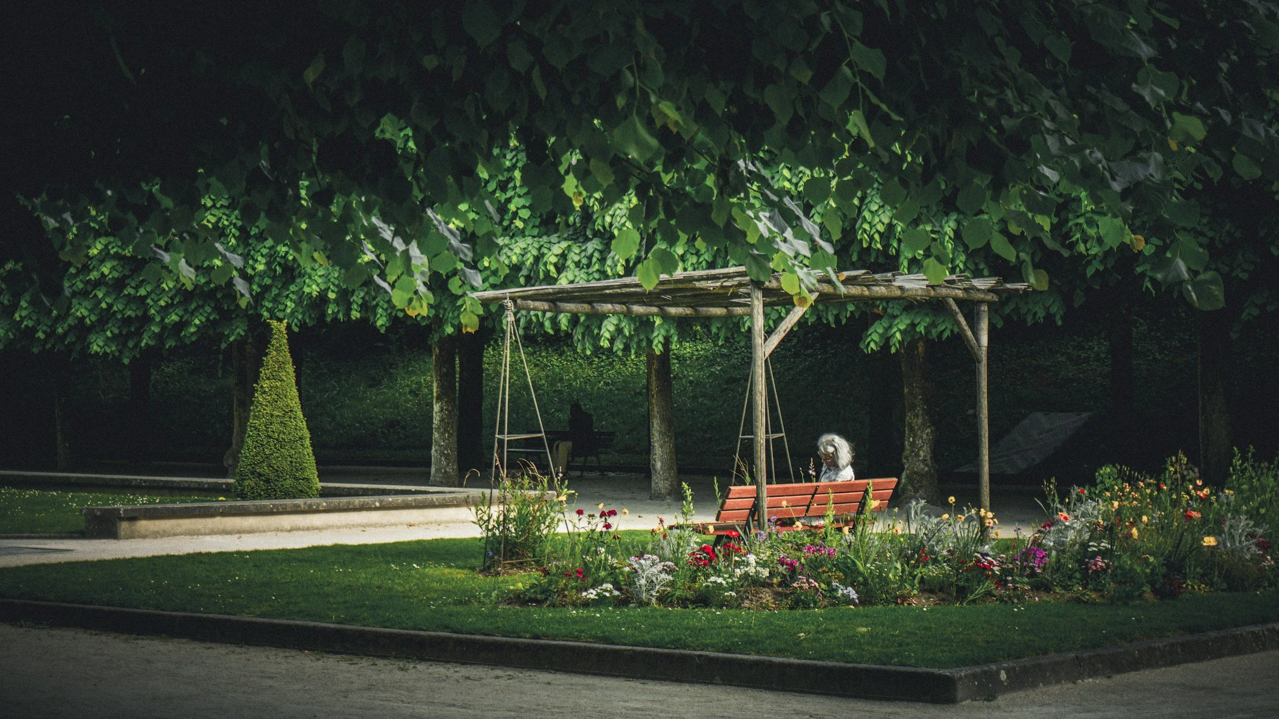A flower bed with various colorful flowers, a wooden bench with a person sitting on it, a wooden swing, and a lush green bush canopy overhead in a park