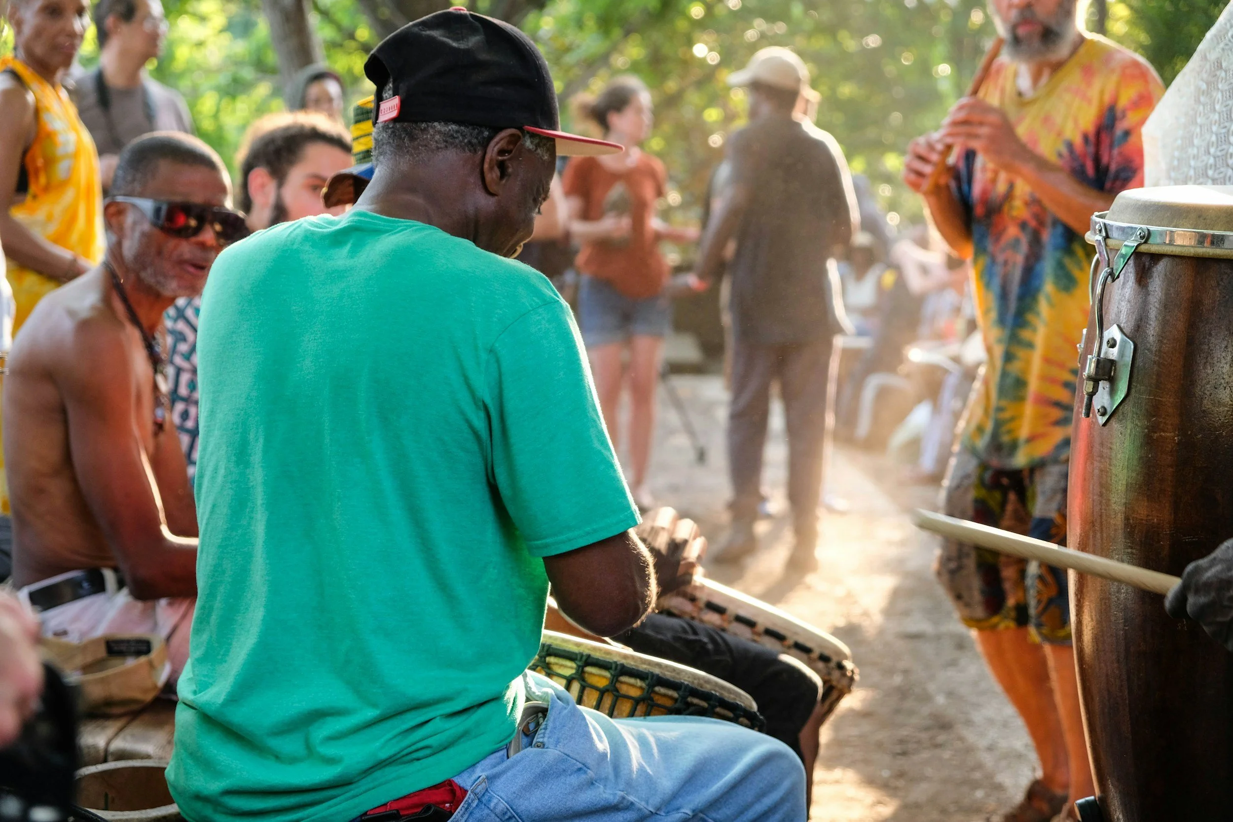 A group of people gathered outdoors, some playing drums and others standing or watching, with the focus on a man in a turquoise shirt and black cap playing a drum. The scene appears to be a lively, sunny event or gathering.