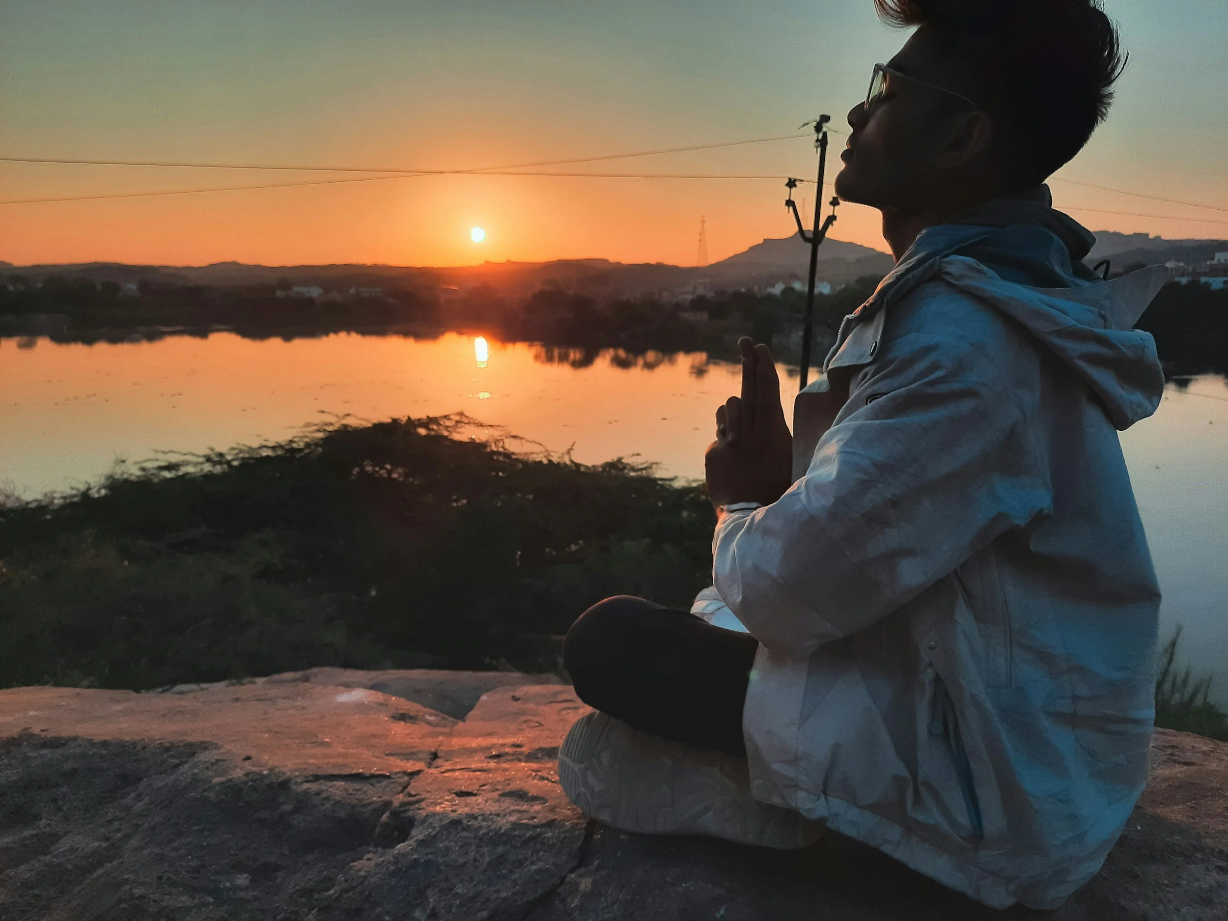 A person sitting cross-legged on a rock, practicing meditation with hands in prayer position, overlooking a river at sunset.