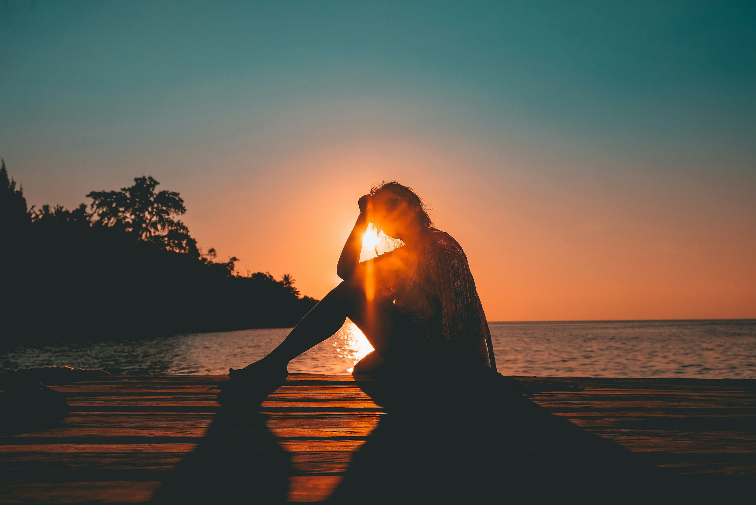 A woman sitting on a dock at sunset, with the sun behind her, creating a silhouette effect.