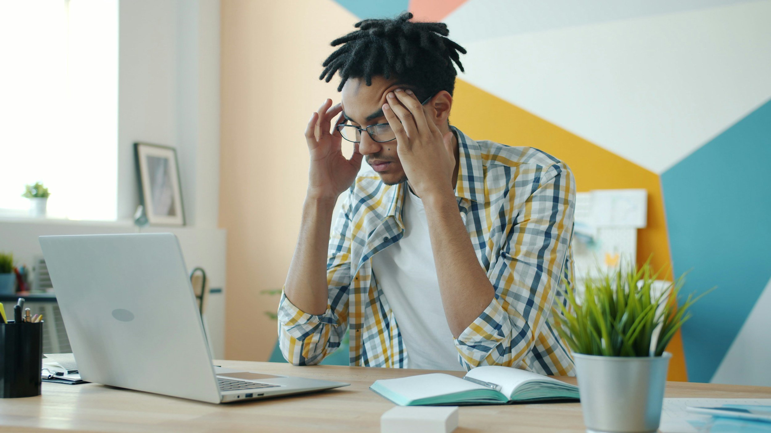 Young man with glasses holding his forehead in frustration at a cluttered workspace with a laptop, open notebook, and a potted plant on the desk.