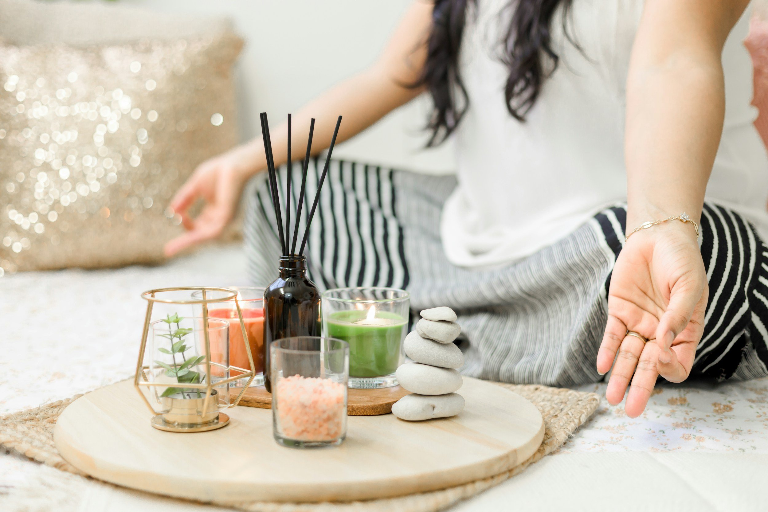 Person sitting on the floor with a wooden tray holding candles, stones, and decorative objects for a home spa or meditation setup.