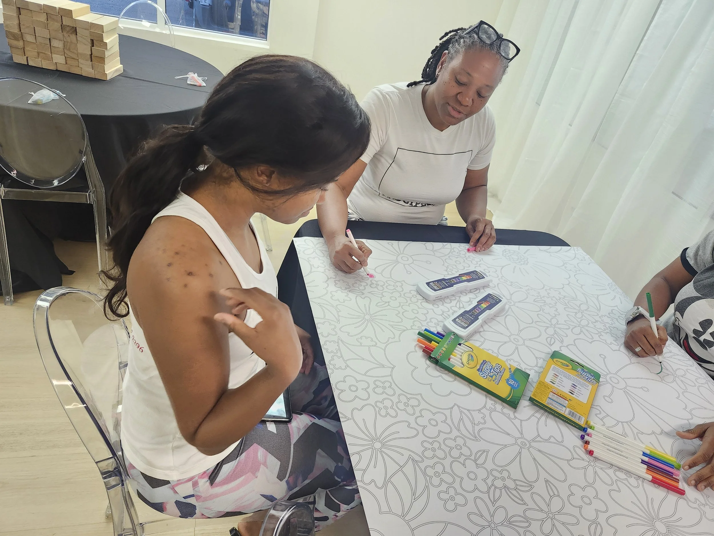 Three women sitting around a table, coloring and drawing with colorful markers and crayons. One woman has visible acne on her arm, and the table has coloring supplies on it.