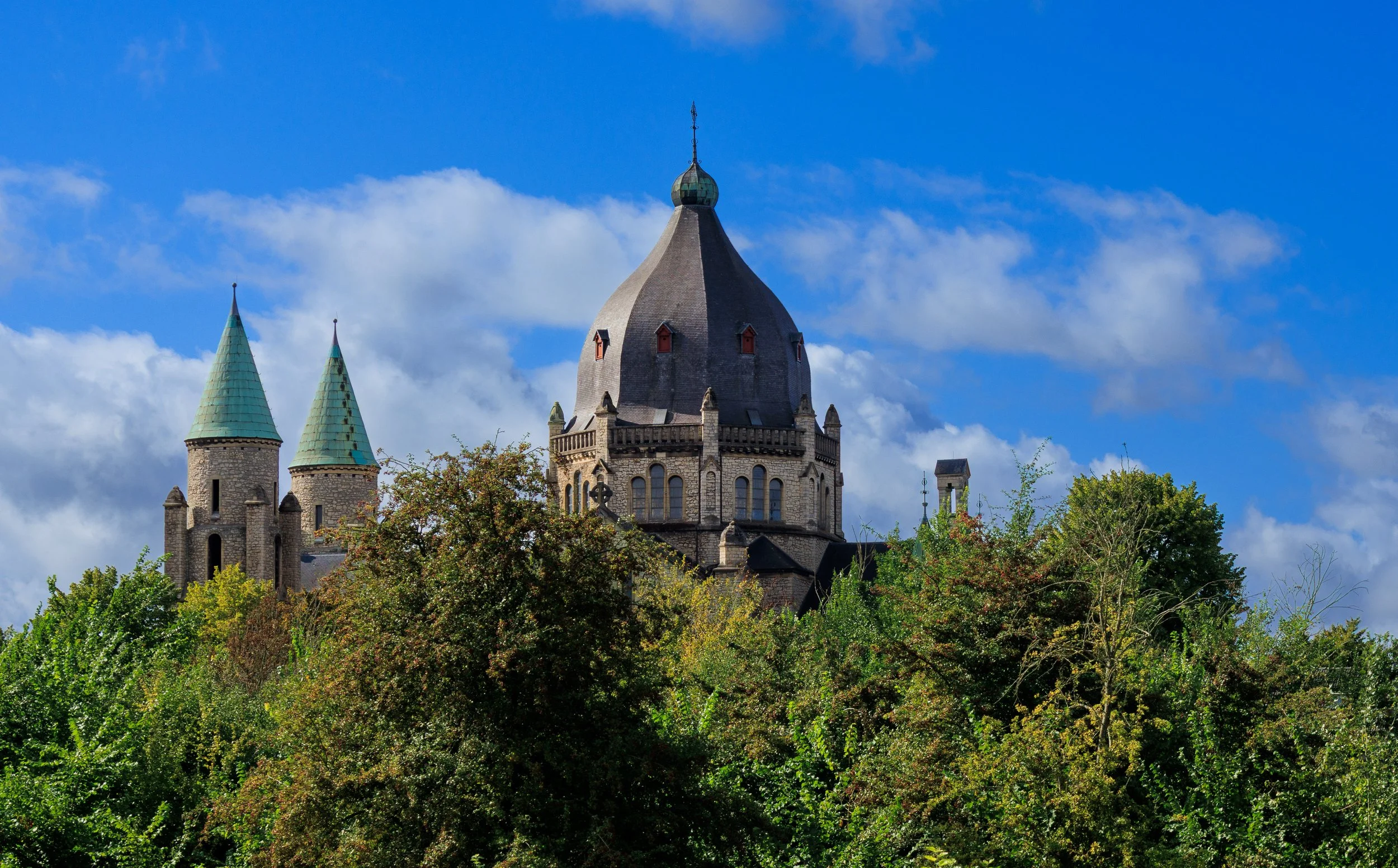 A castle with tall towers and a large central dome, surrounded by green trees, under a partly cloudy blue sky.