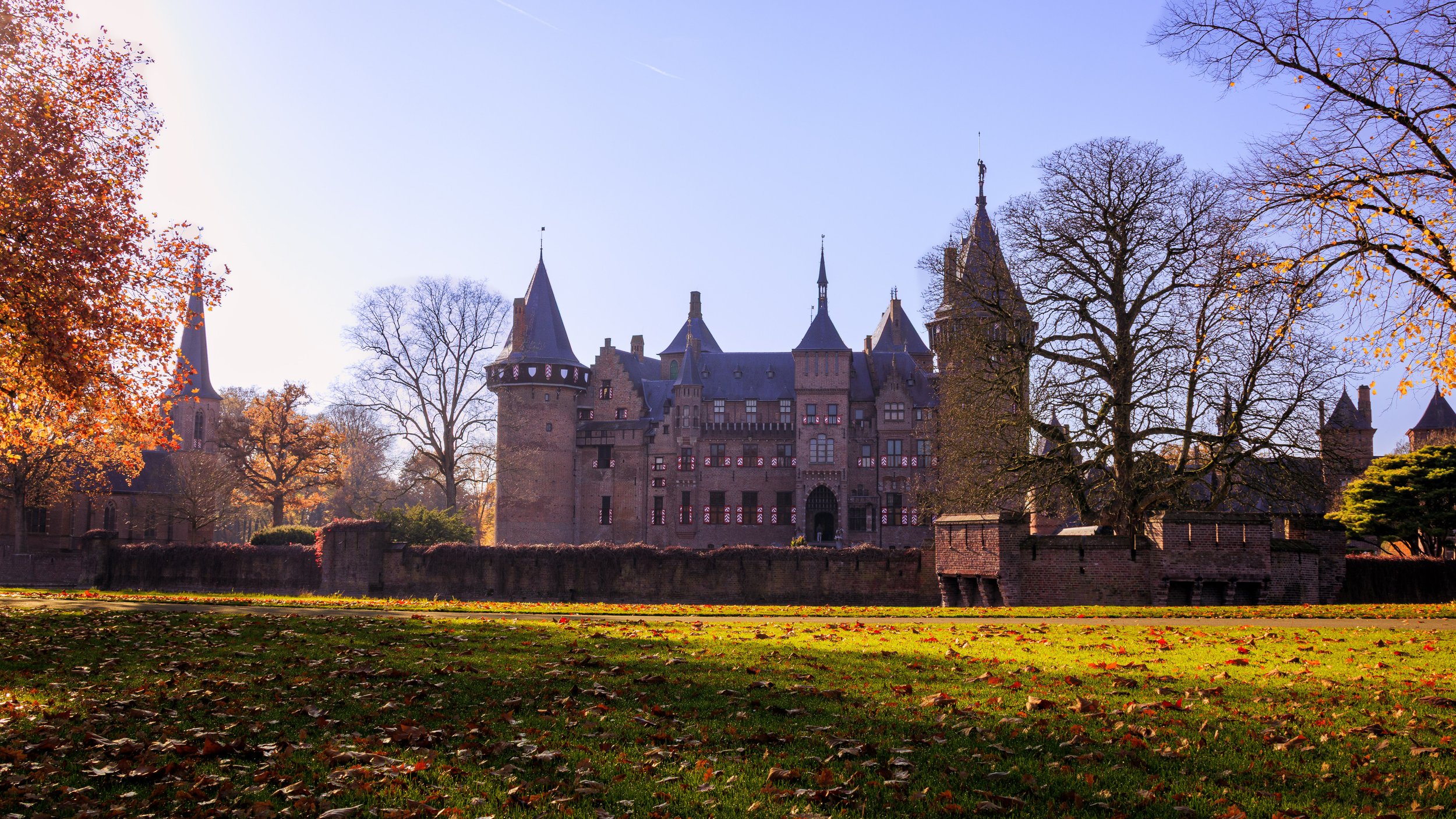A large medieval castle with towers and turrets, surrounded by autumn trees and fallen leaves on the grass.