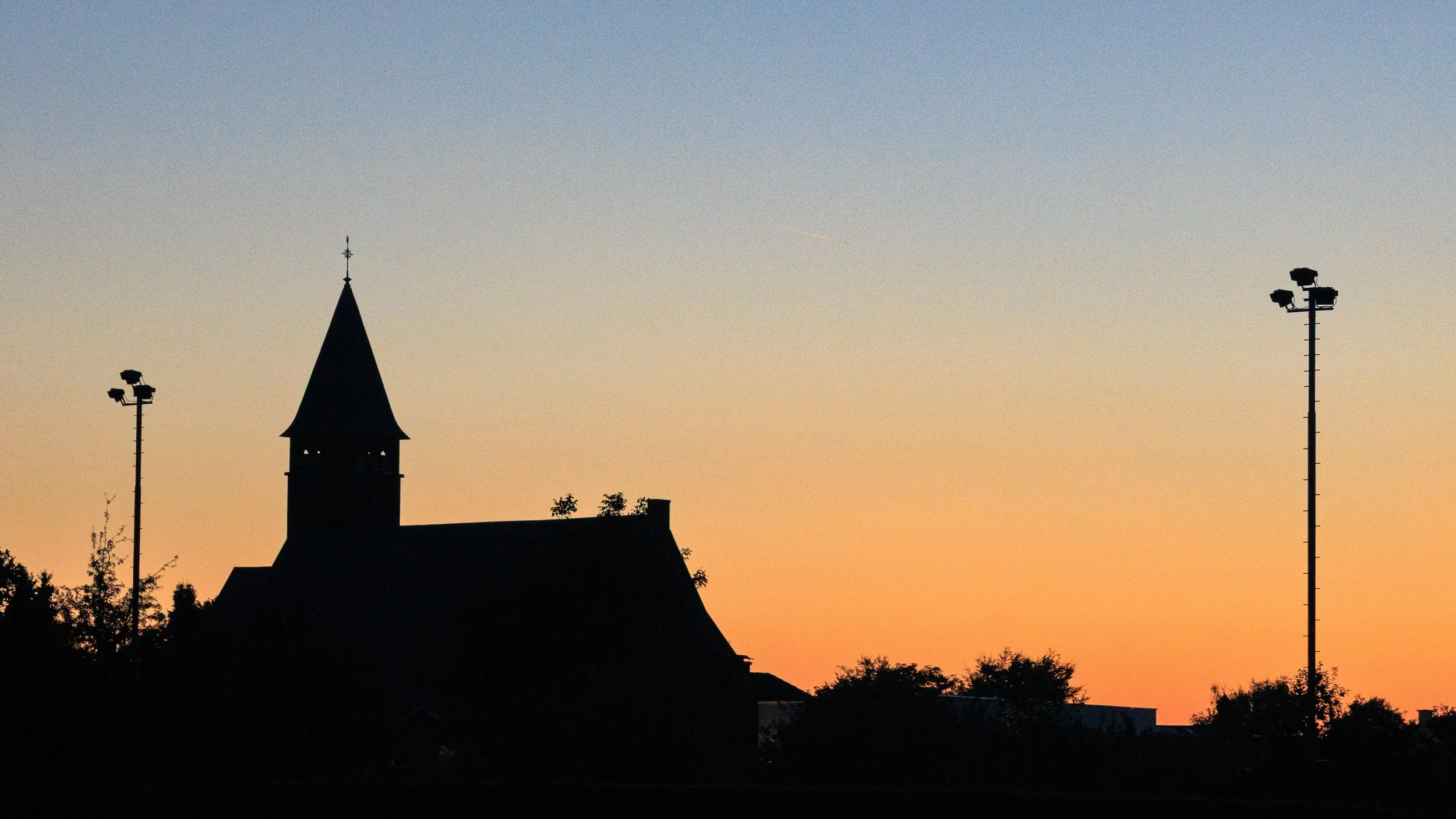 Silhouette of a church with a steeple, two tall light poles, and trees against a sunset sky with orange and blue hues.
