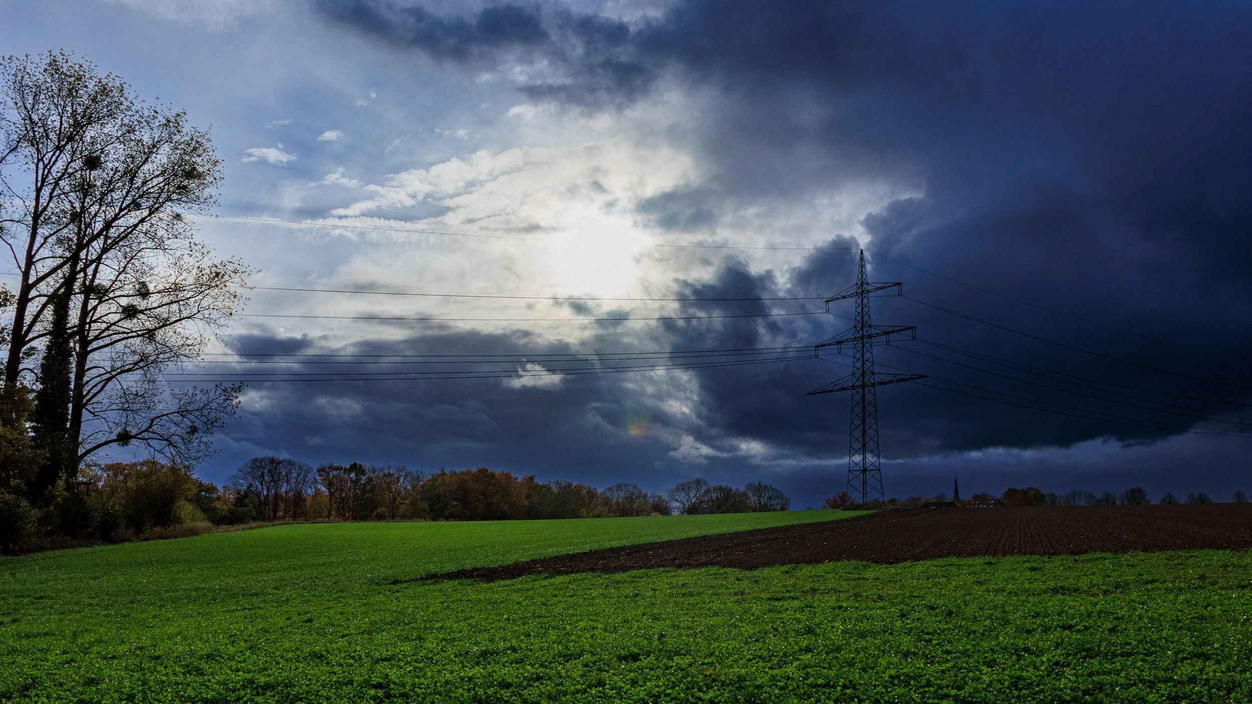 A landscape of green fields with trees on the left and in the background, under a dramatic cloudy sky with the sun obscured, and power lines and a transmission tower crossing the scene.