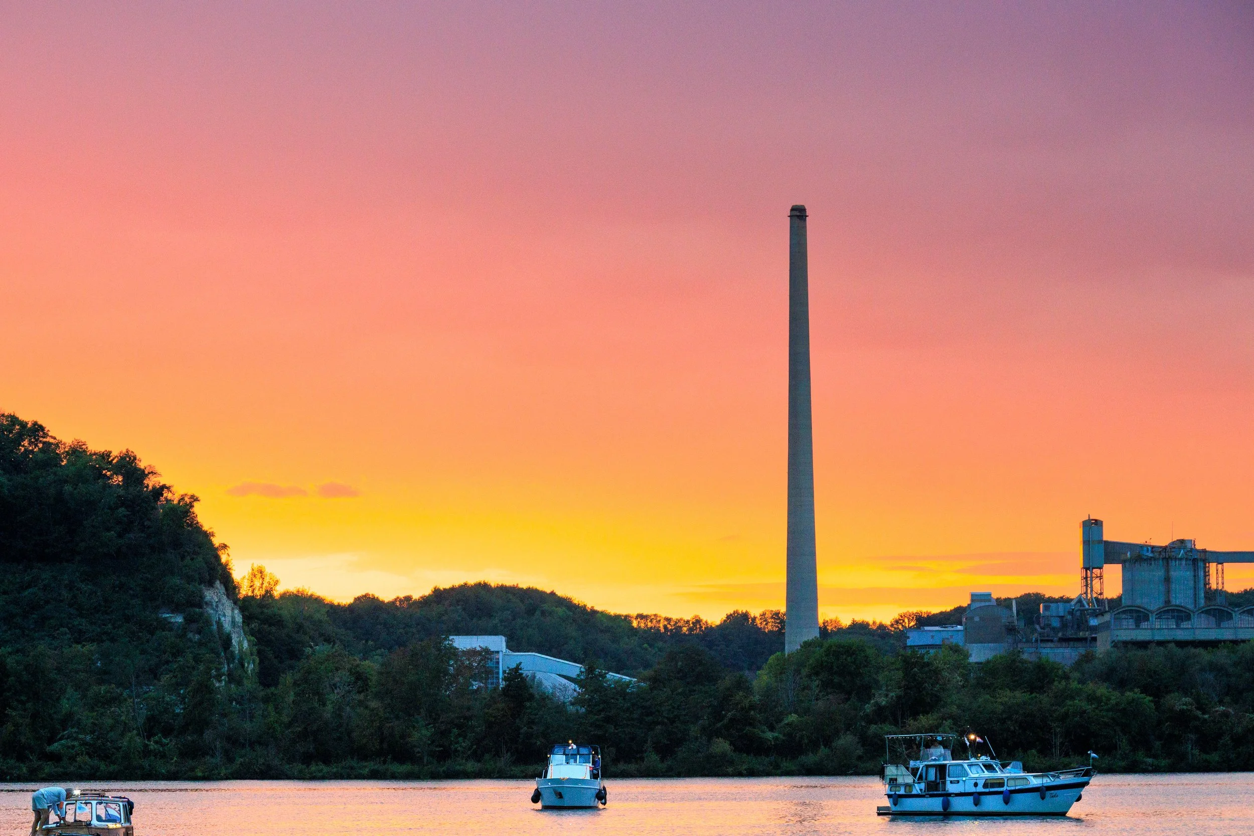 Colorful sunset over a river with boats, industrial buildings, and a tall smokestack.