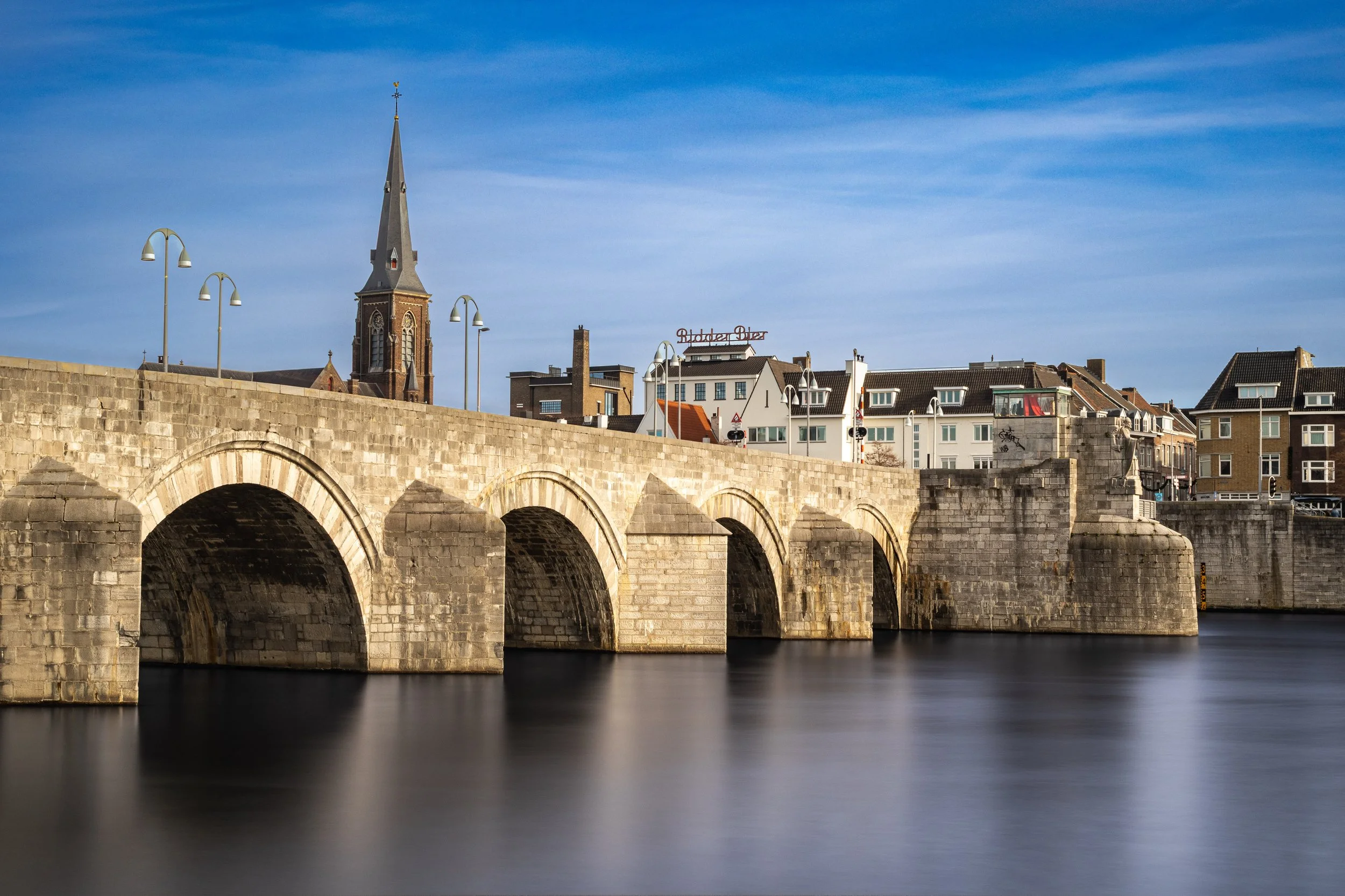 A stone bridge over calm water with a city skyline in the background, including a church steeple and various buildings, under a blue sky.