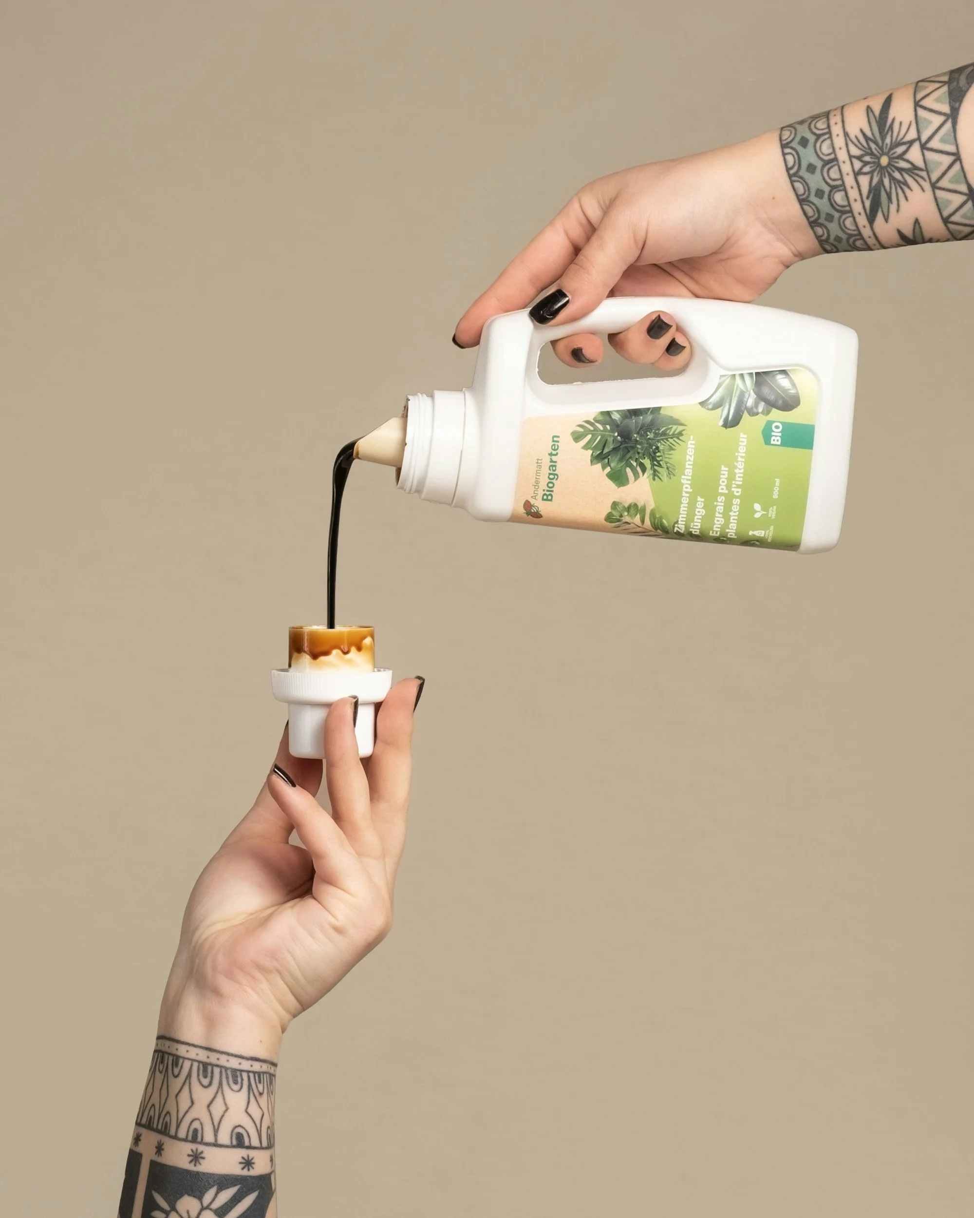 Hand pouring brown liquid from a white bottle onto a small white cup held by another hand with tattoos, against a plain beige background.