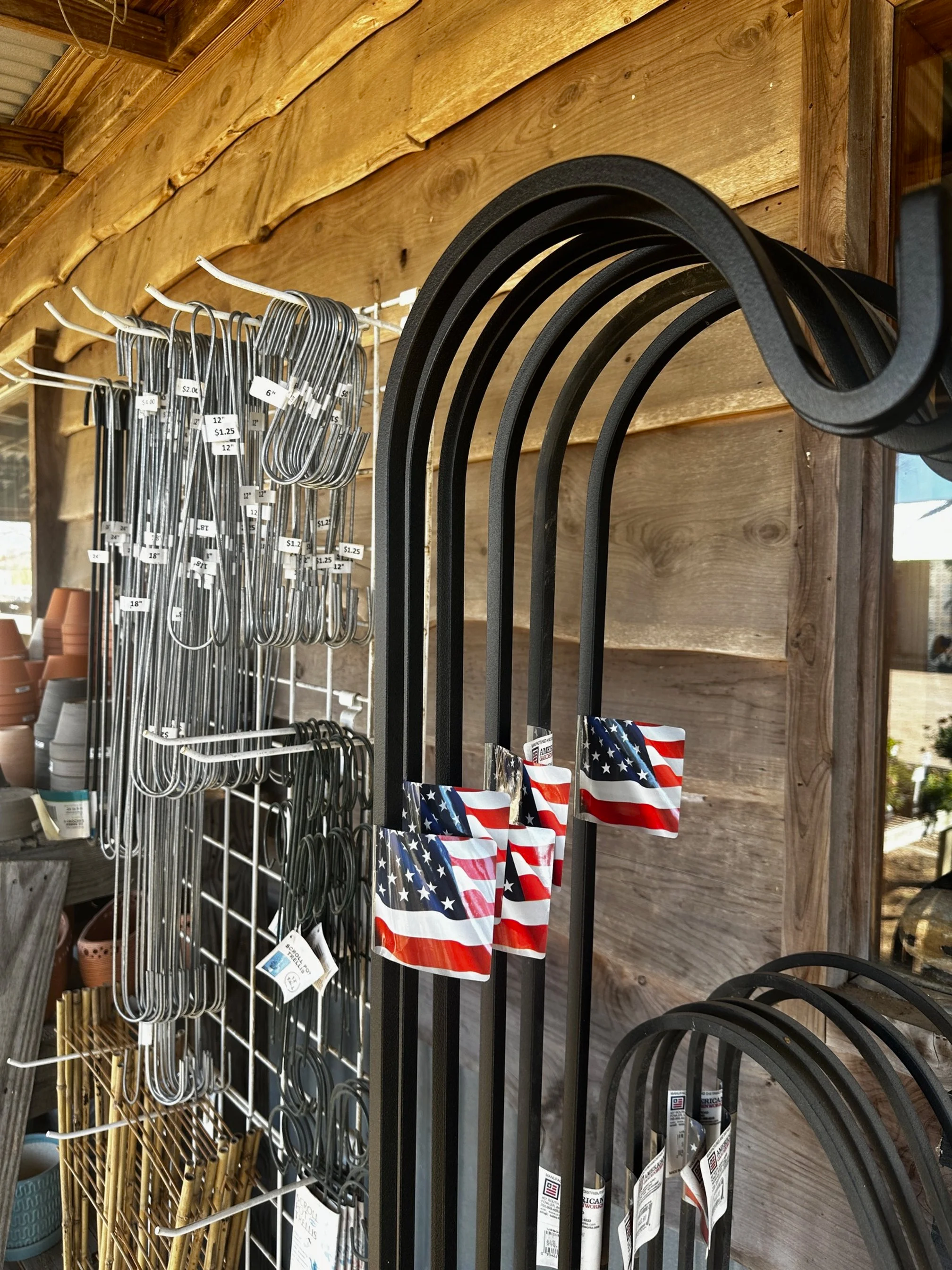Black metal outdoor supports and a display of galvanized metal hooks and garden tools with American flag tags at a garden store.