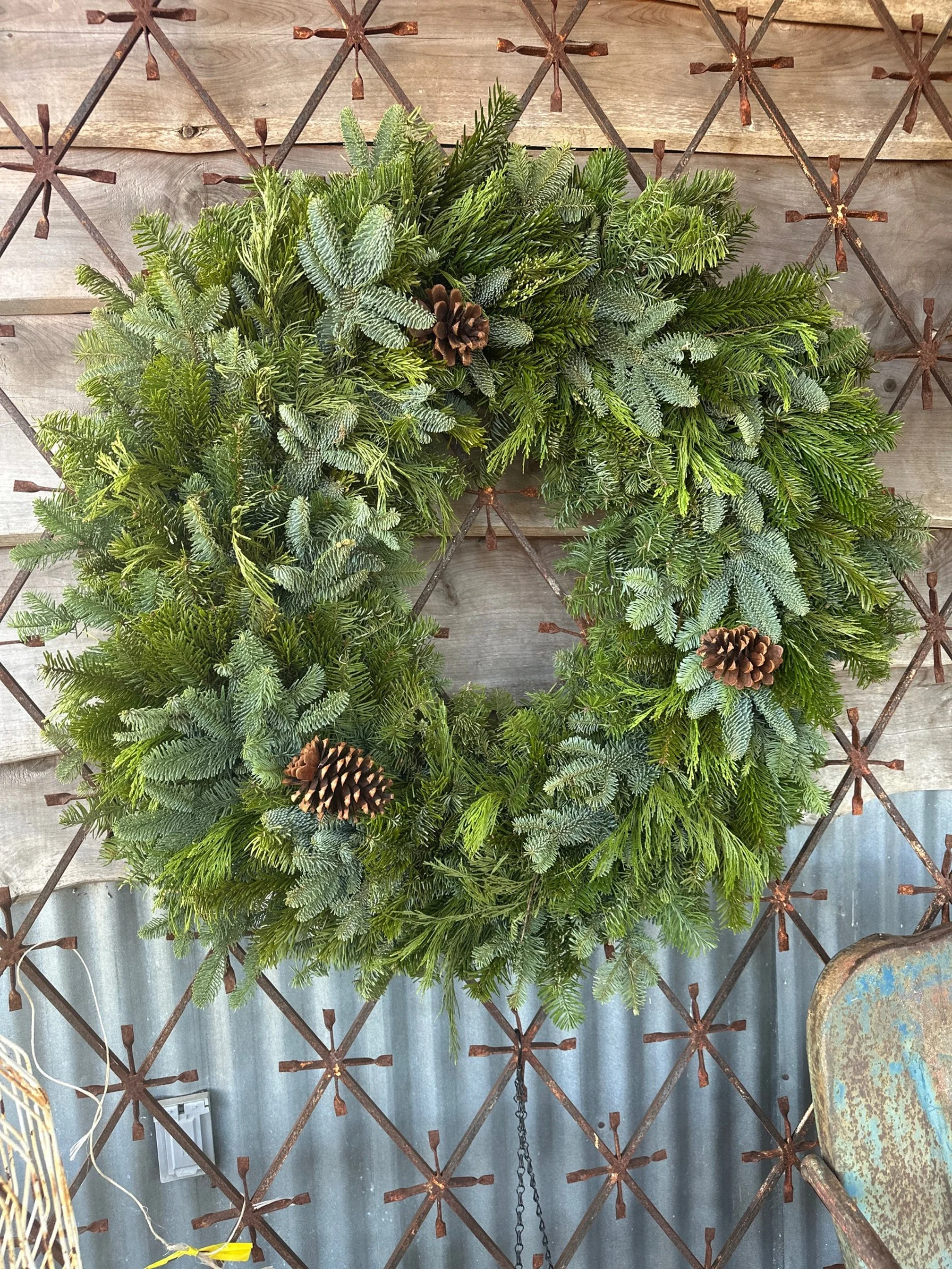 A decorative Christmas wreath made of green pine and fir branches, decorated with three brown pinecones, hanging against a rustic wooden and corrugated metal wall.