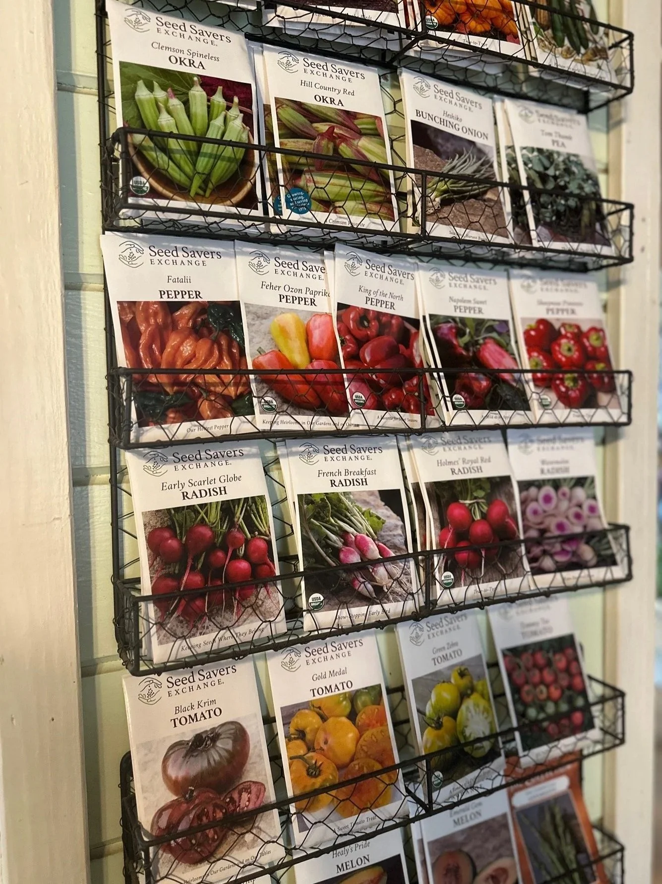 Display of seed packets for various vegetables and flowers on wire racks, including okra, pepper, radish, tomato, melon, and other seeds.